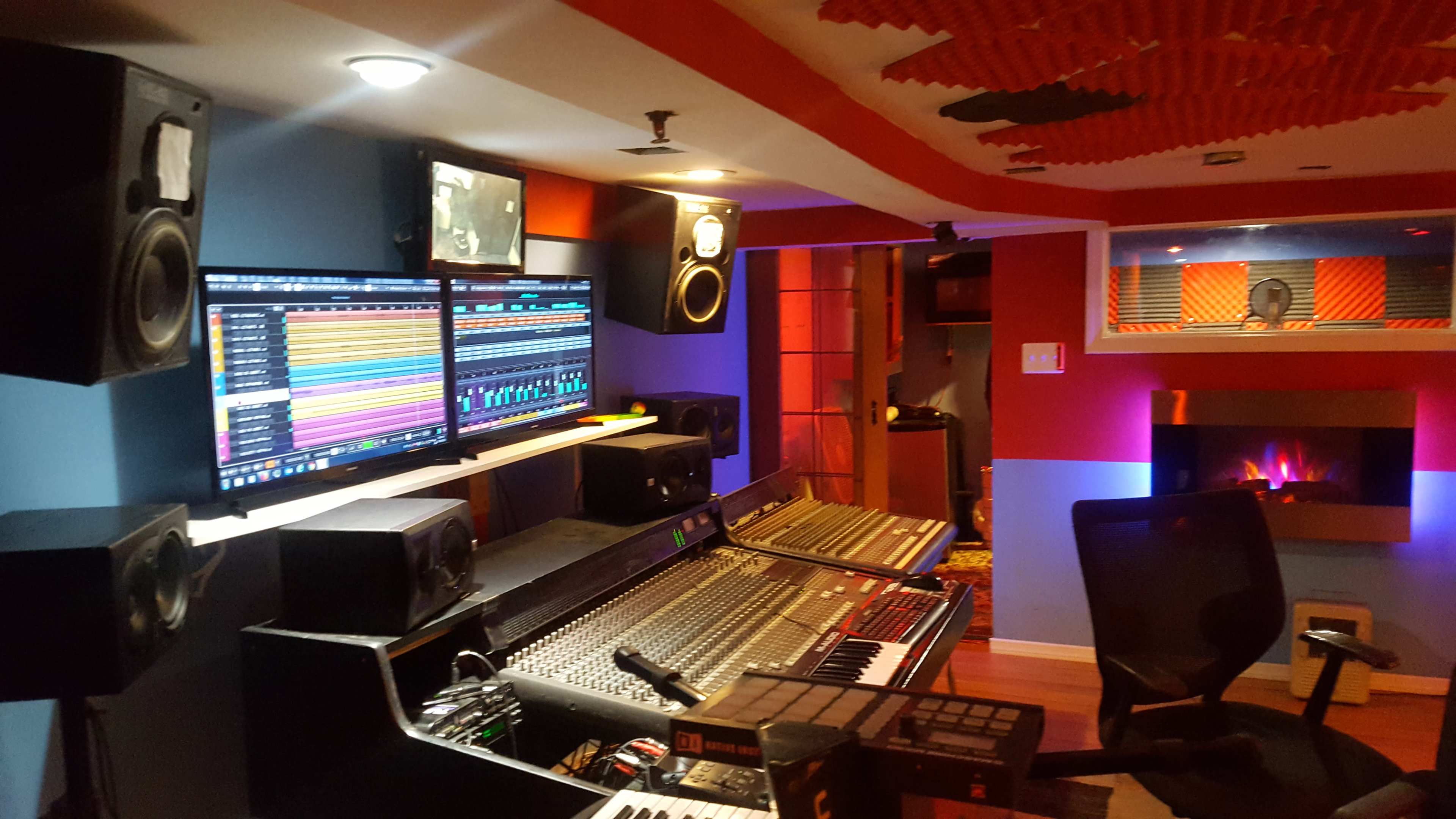 A modern recording studio with a mixing console, dual computer monitors displaying audio tracks, and speakers arranged around a slightly dimly lit room.