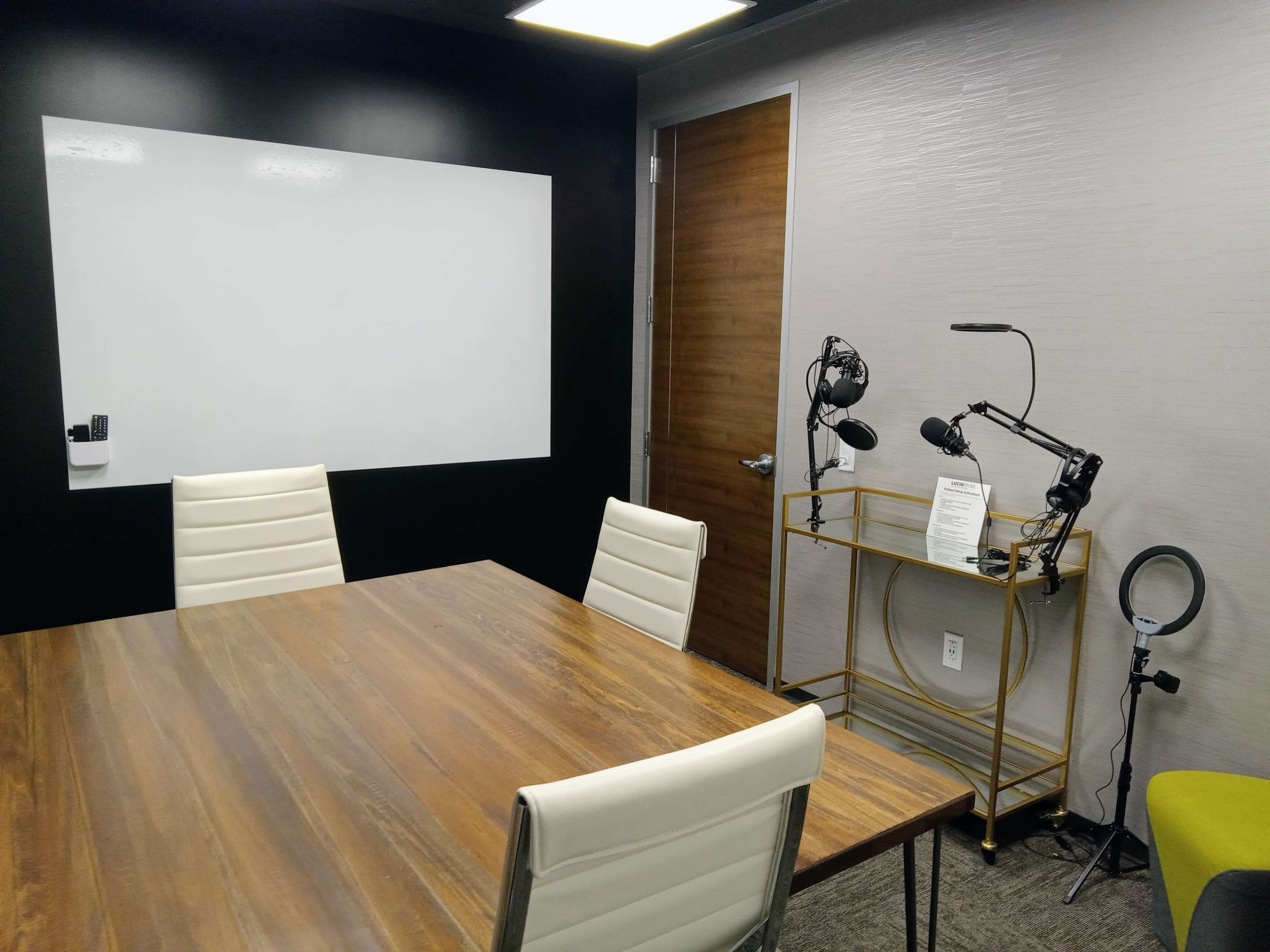 A conference room features a wooden table, white chairs, a large whiteboard, and a standing cart with microphones and a ring light.
