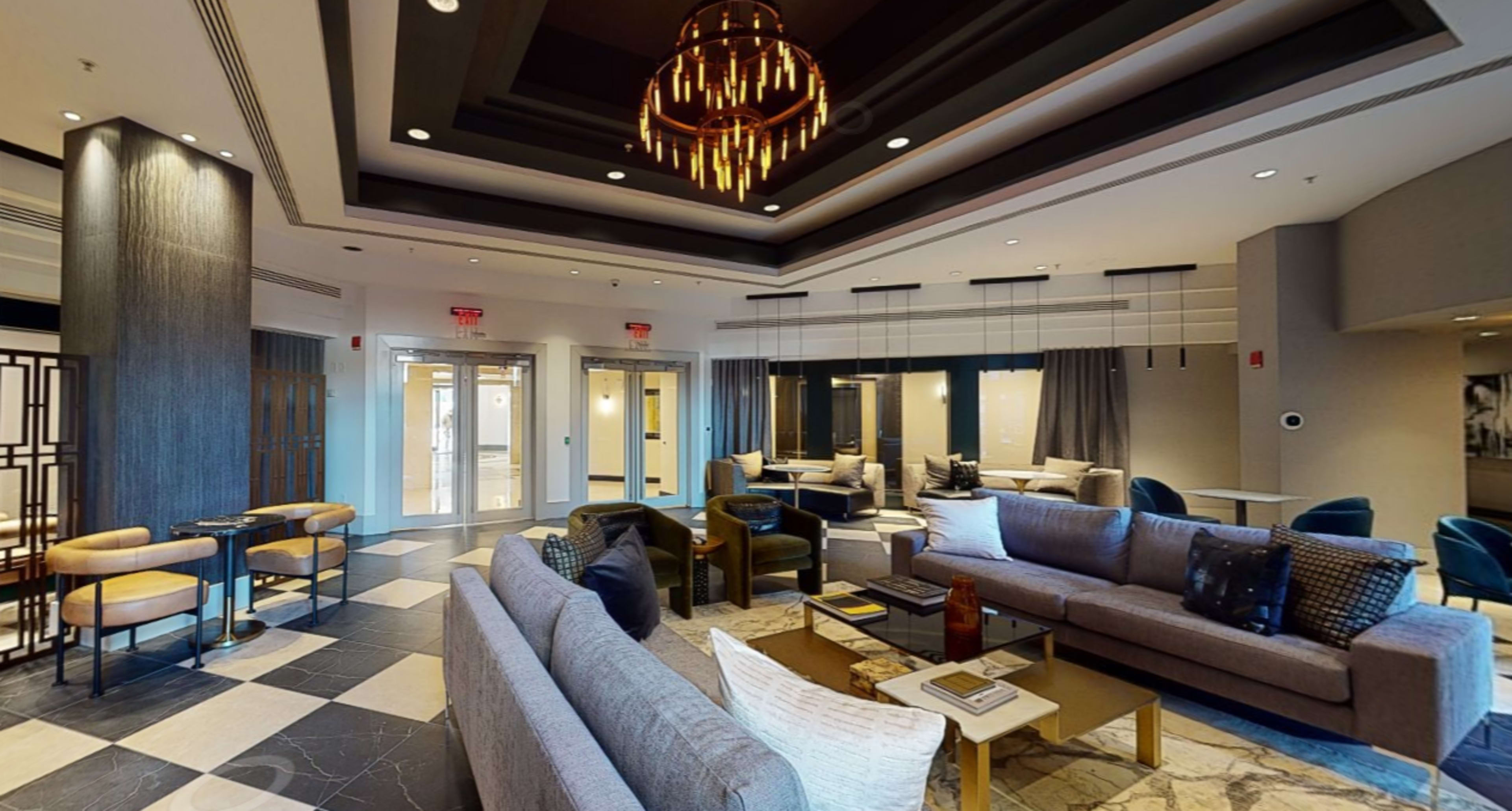 A modern lobby with a seating area featuring gray sofas, a chandelier, and large windows.