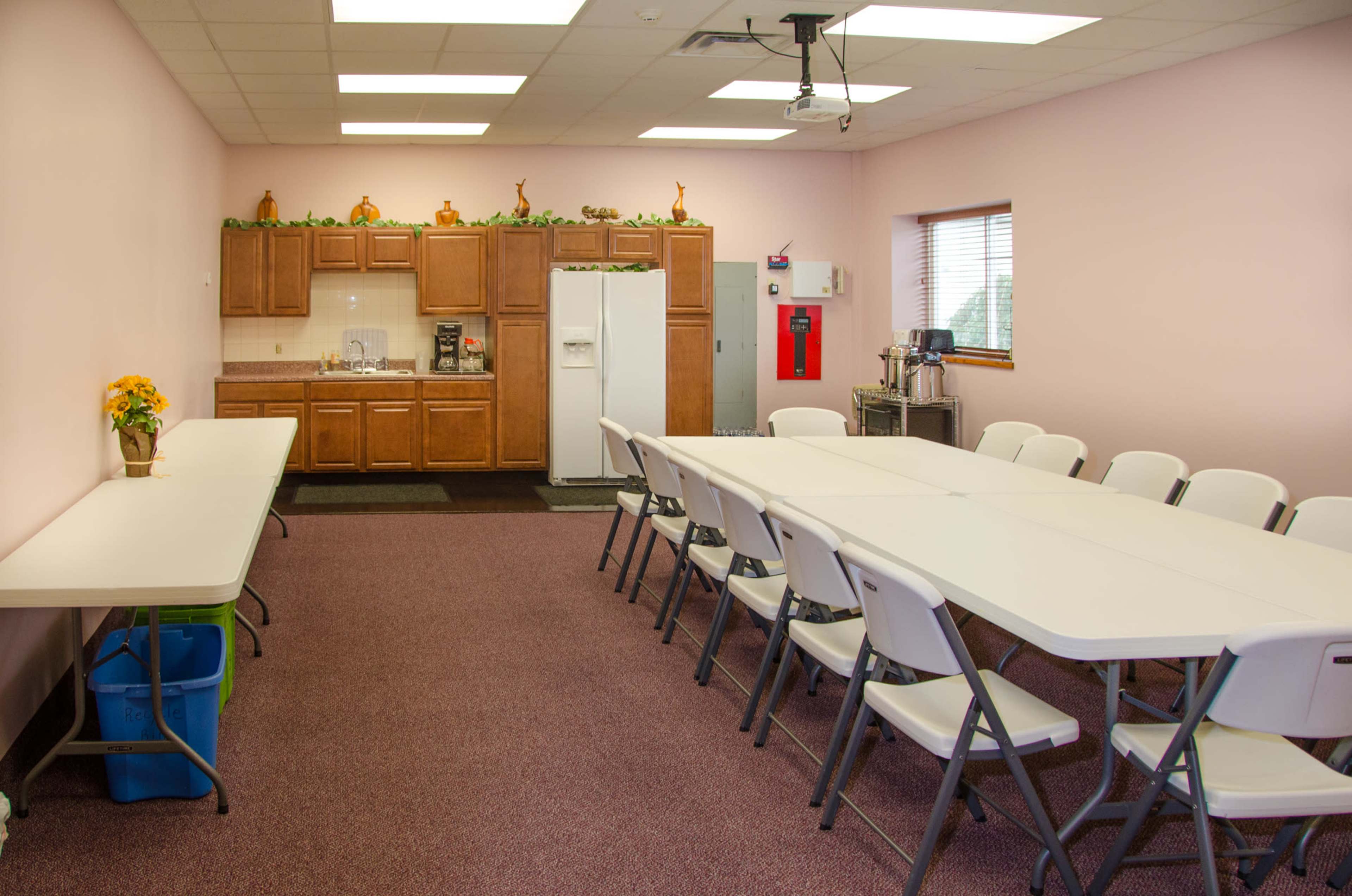 A large room with a long table surrounded by folding chairs, a kitchenette with wooden cabinets, and a small area for beverages.