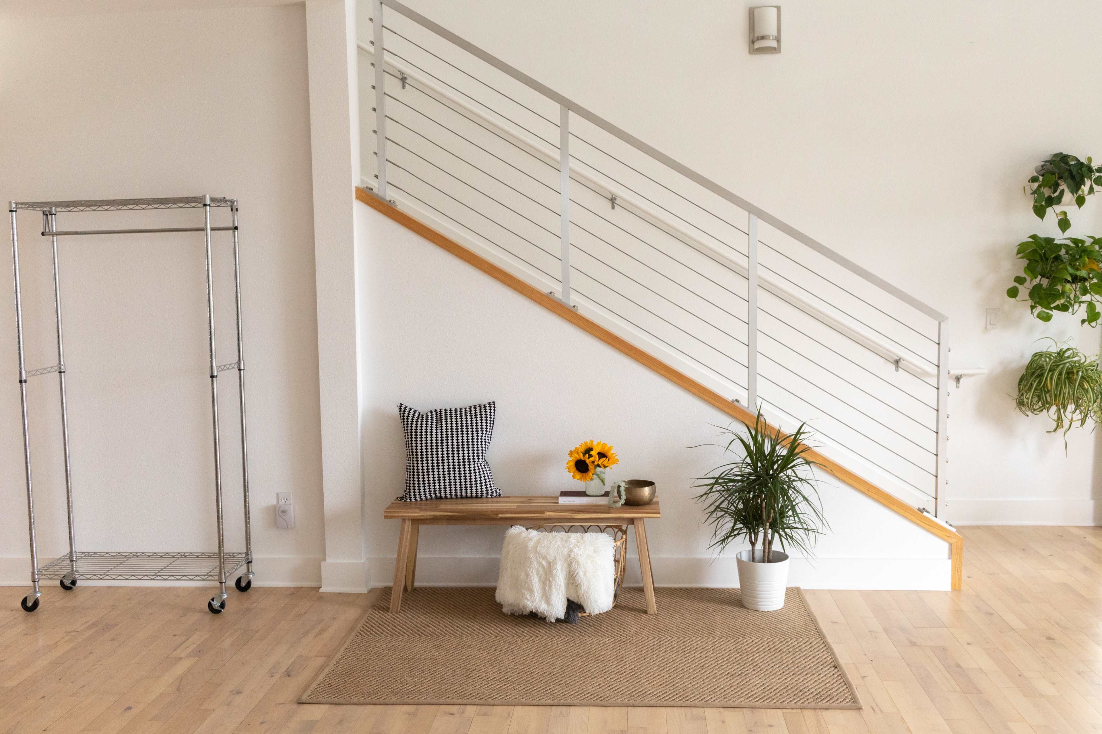 A minimalist interior features a wooden bench with a plant and a vase of sunflowers against a wall with a staircase and a clothing rack nearby.