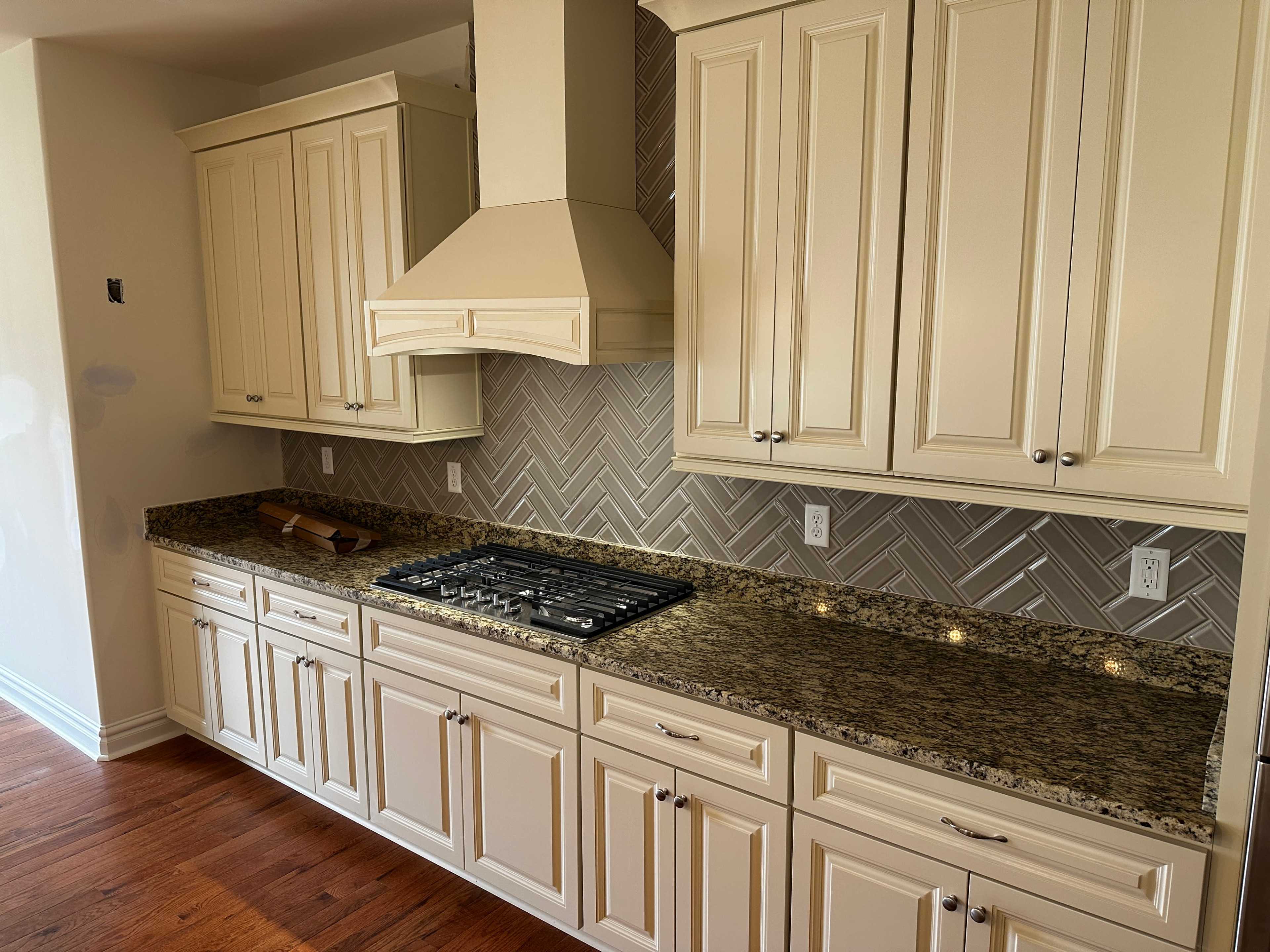 A kitchen with light-colored cabinetry and a granite countertop, featuring a gas stove and a herringbone-patterned tile backsplash.