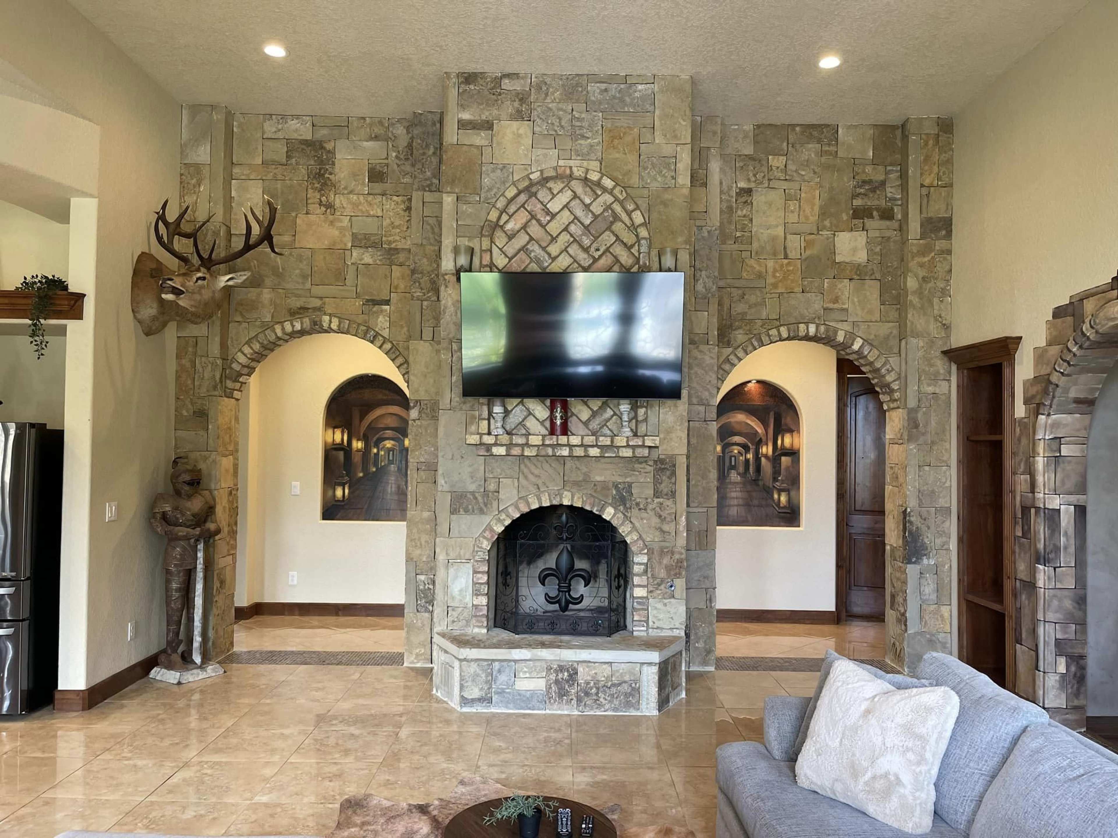 The interior of a spacious living room features a stone accent wall with a large TV, a fireplace, and arched doorways leading to other rooms.