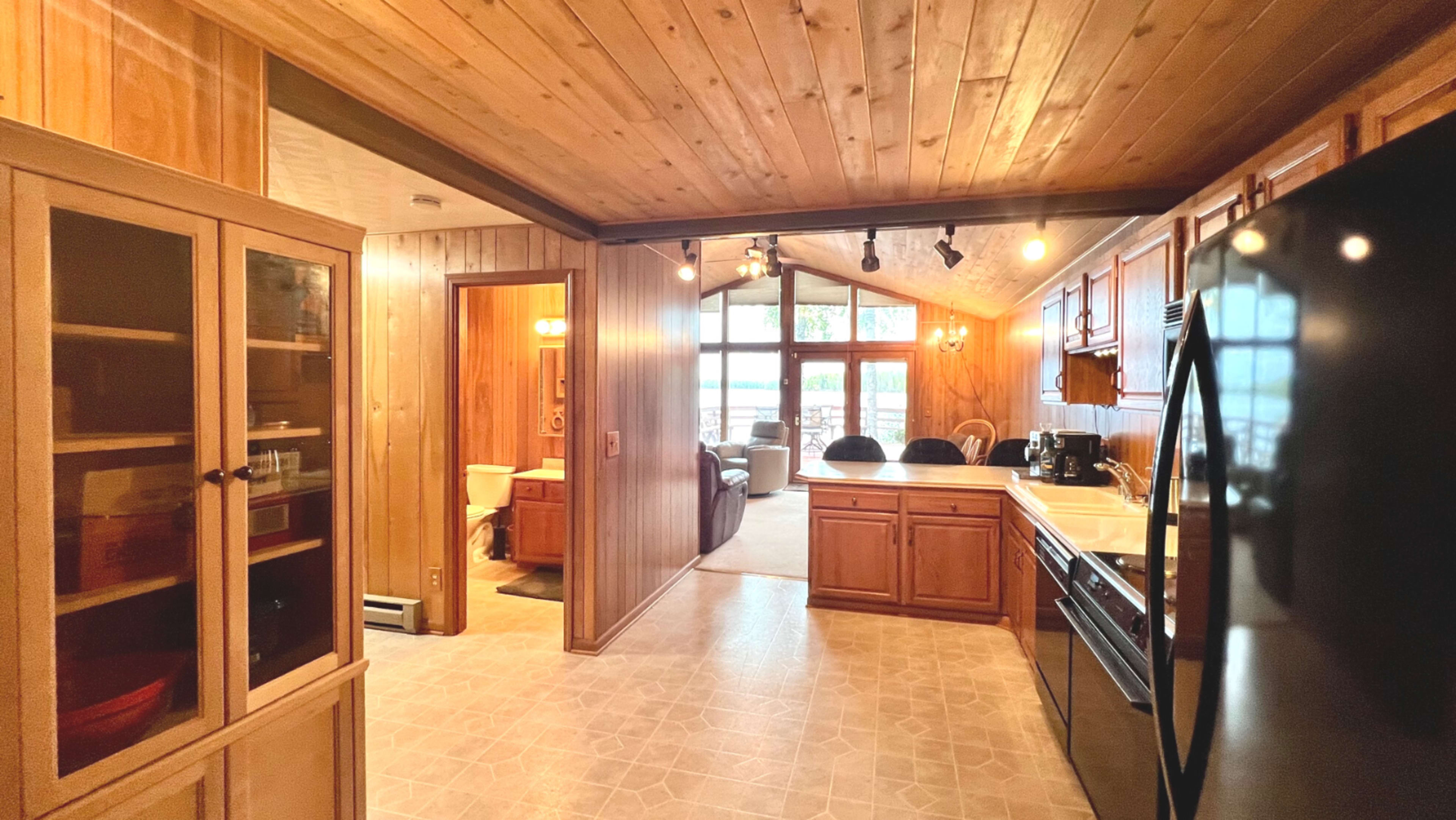 A wooden-finished kitchen with a view into a sunlit living area, featuring a refrigerator, stove, and cabinetry.
