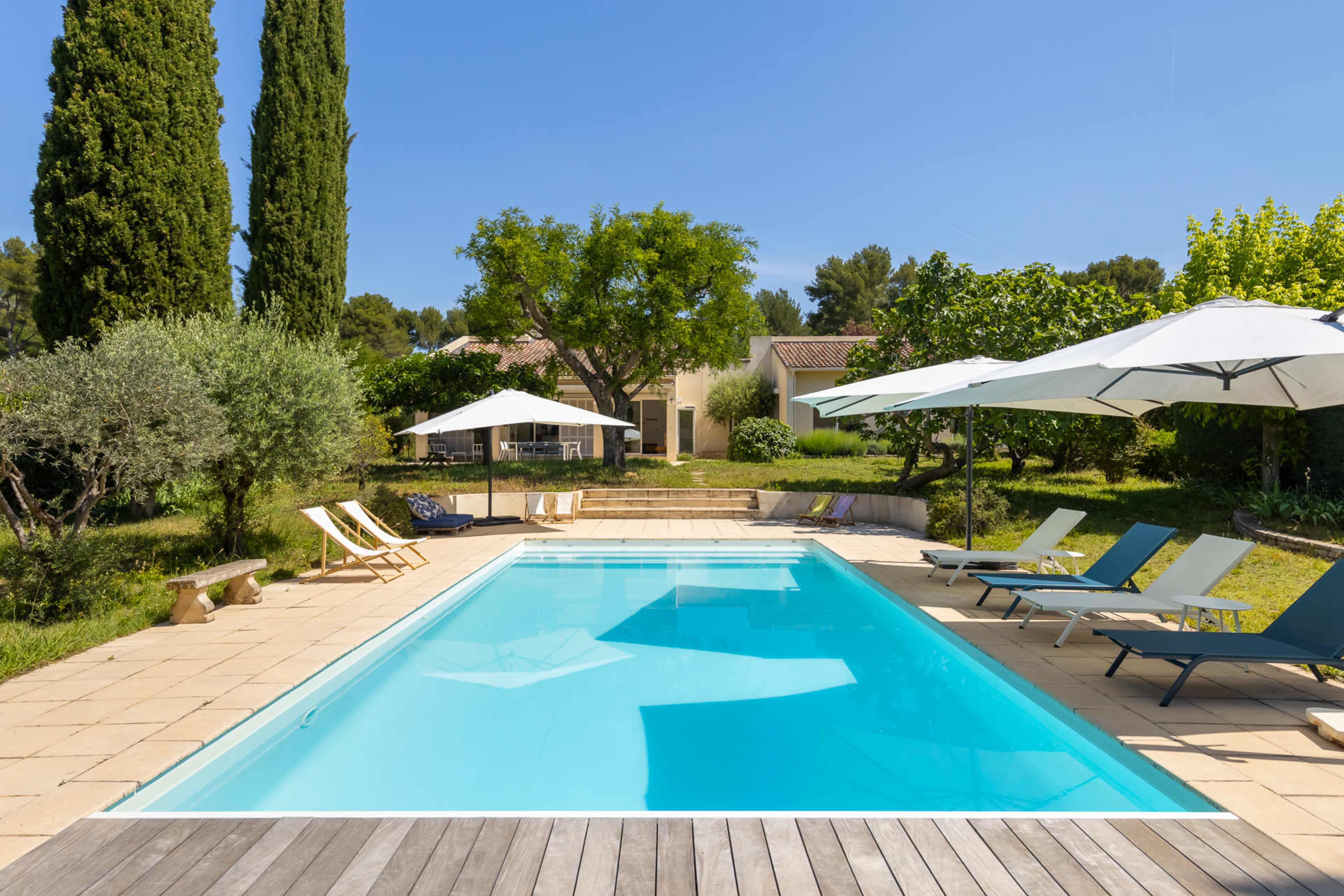 A clear swimming pool is surrounded by lounge chairs and parasols in a landscaped garden with trees and a house in the background.