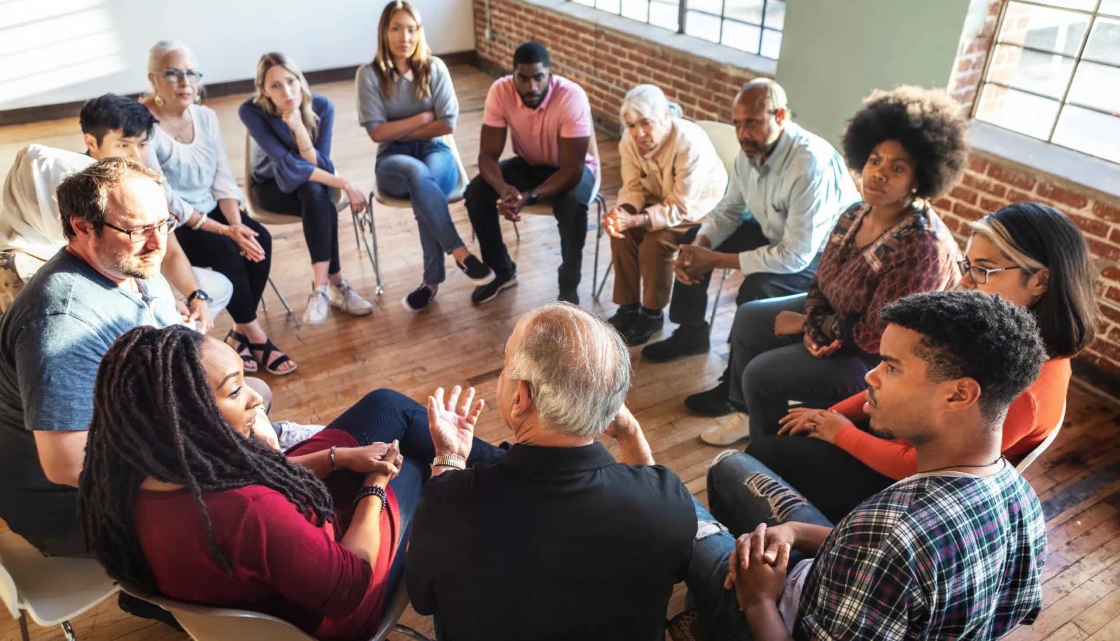A diverse group of individuals sits in a circle, engaging in a discussion in a well-lit room with large windows and exposed brick walls.