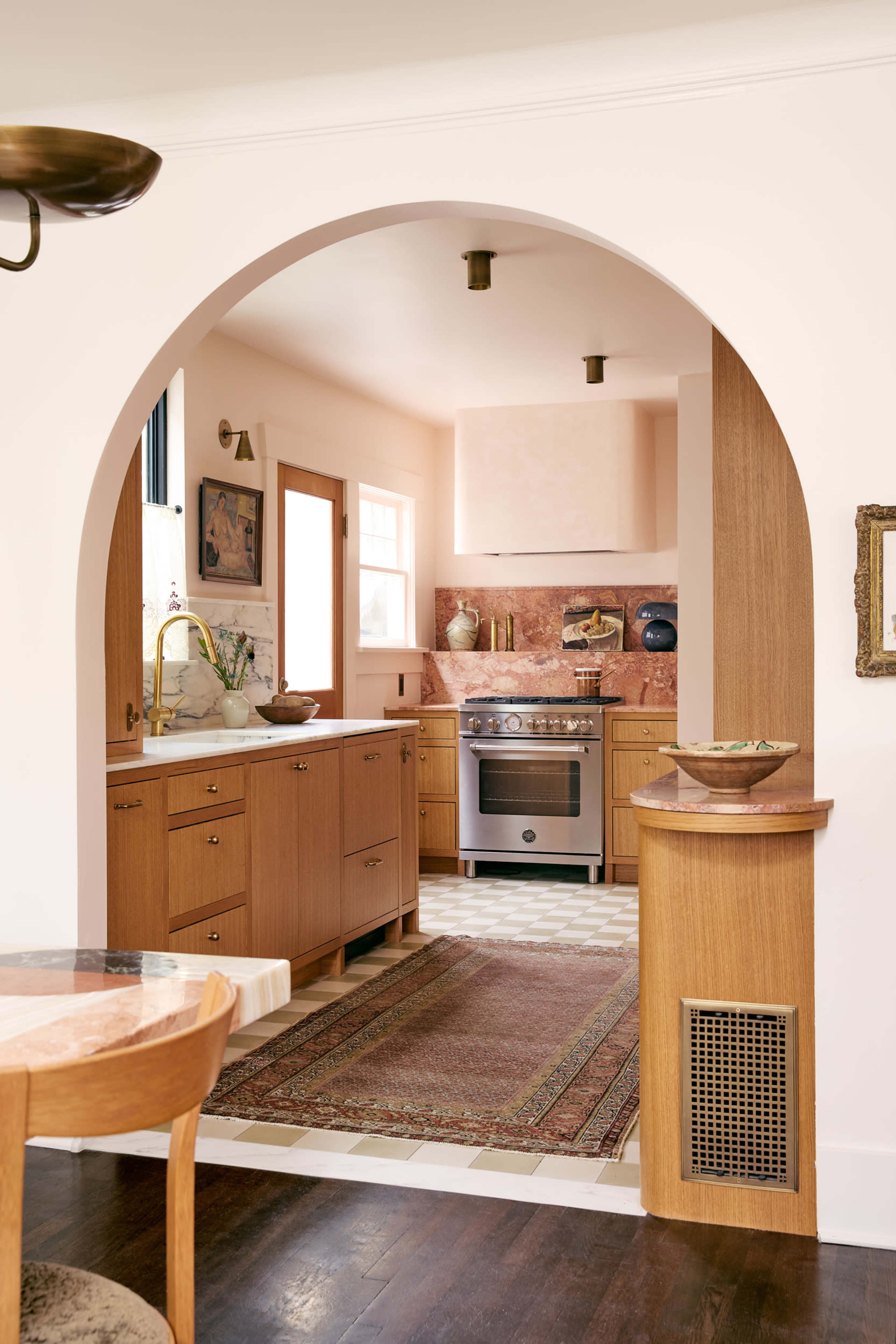 The image depicts a kitchen through an arched opening, featuring wooden cabinets, a stove, and a patterned rug on the floor.