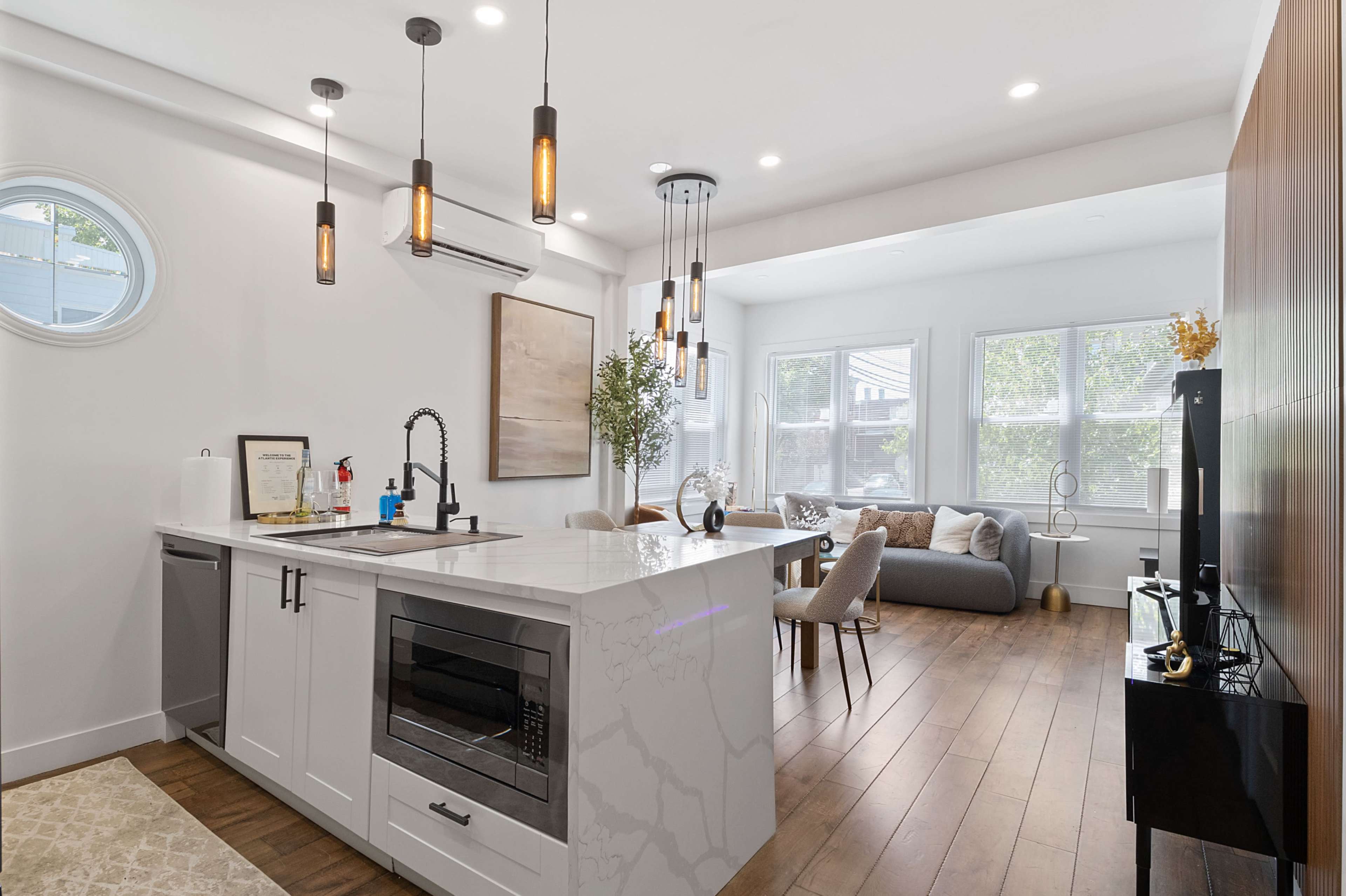 A modern kitchen with a white marble island and pendant lights opens into a bright living area featuring large windows and contemporary furniture.