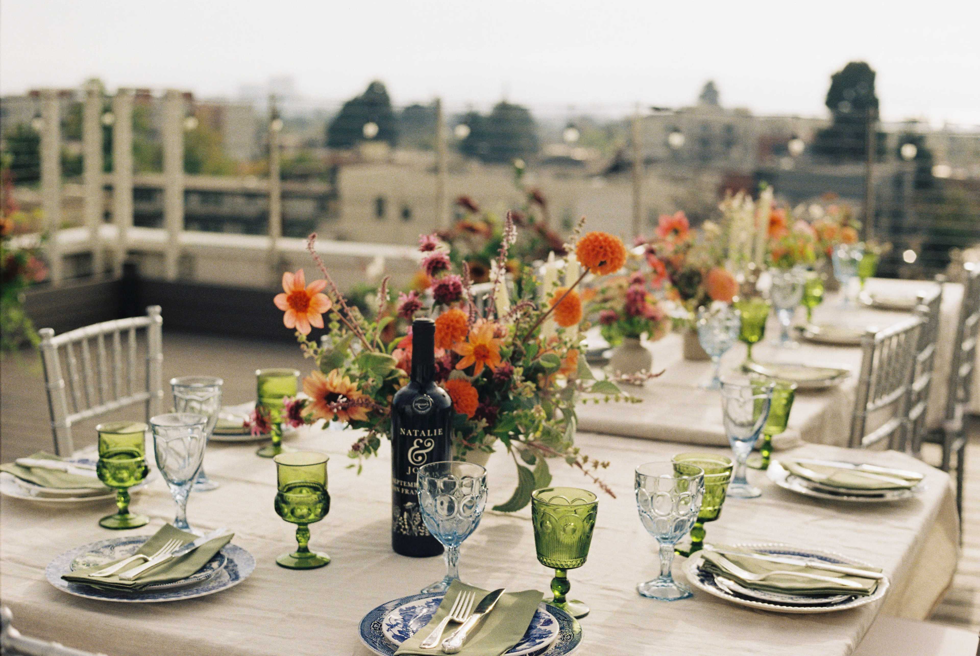 A beautifully arranged outdoor dining table features colorful flowers, green glassware, and a bottle of wine among elegant place settings.
