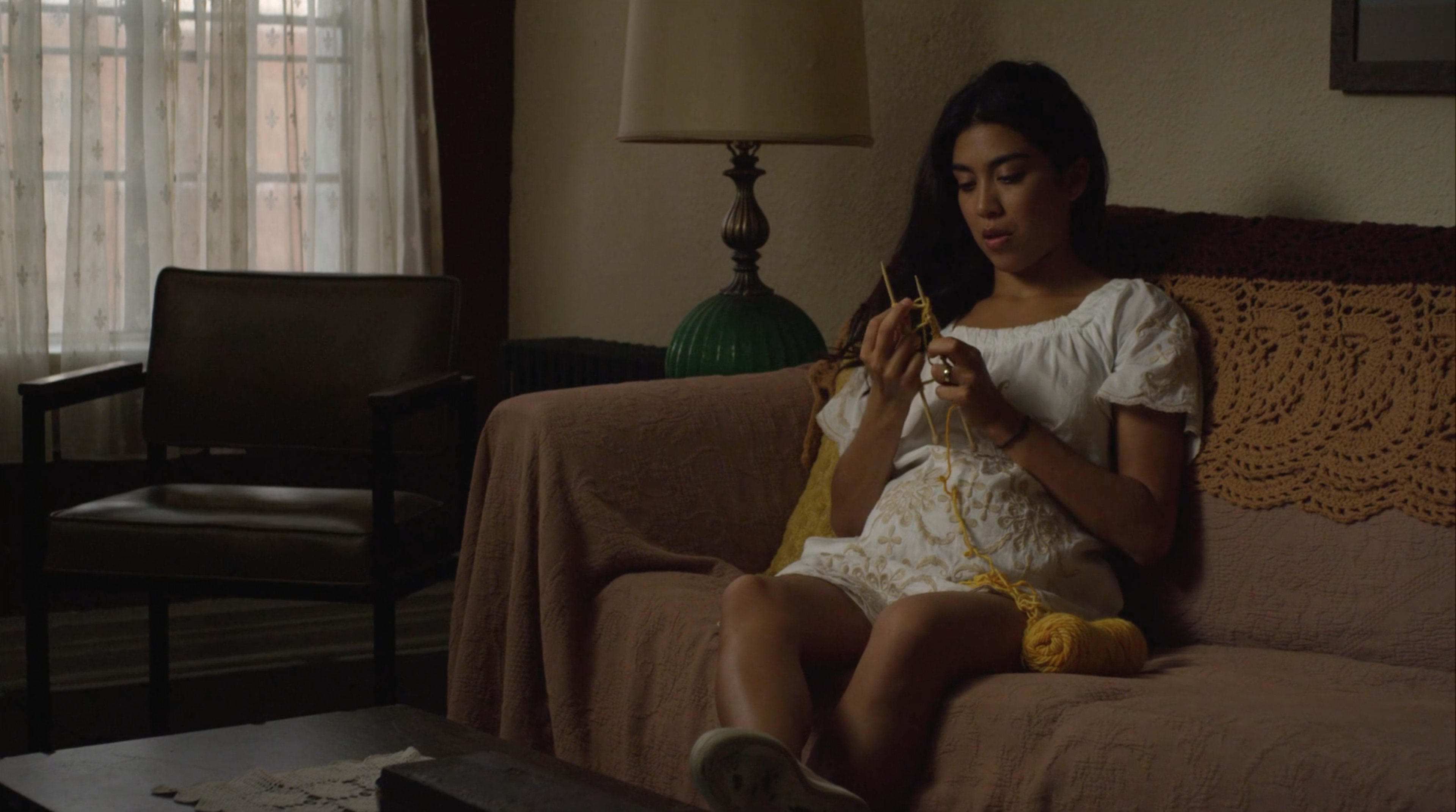 A woman sits on a couch in a well-lit room, knitting with yellow yarn.