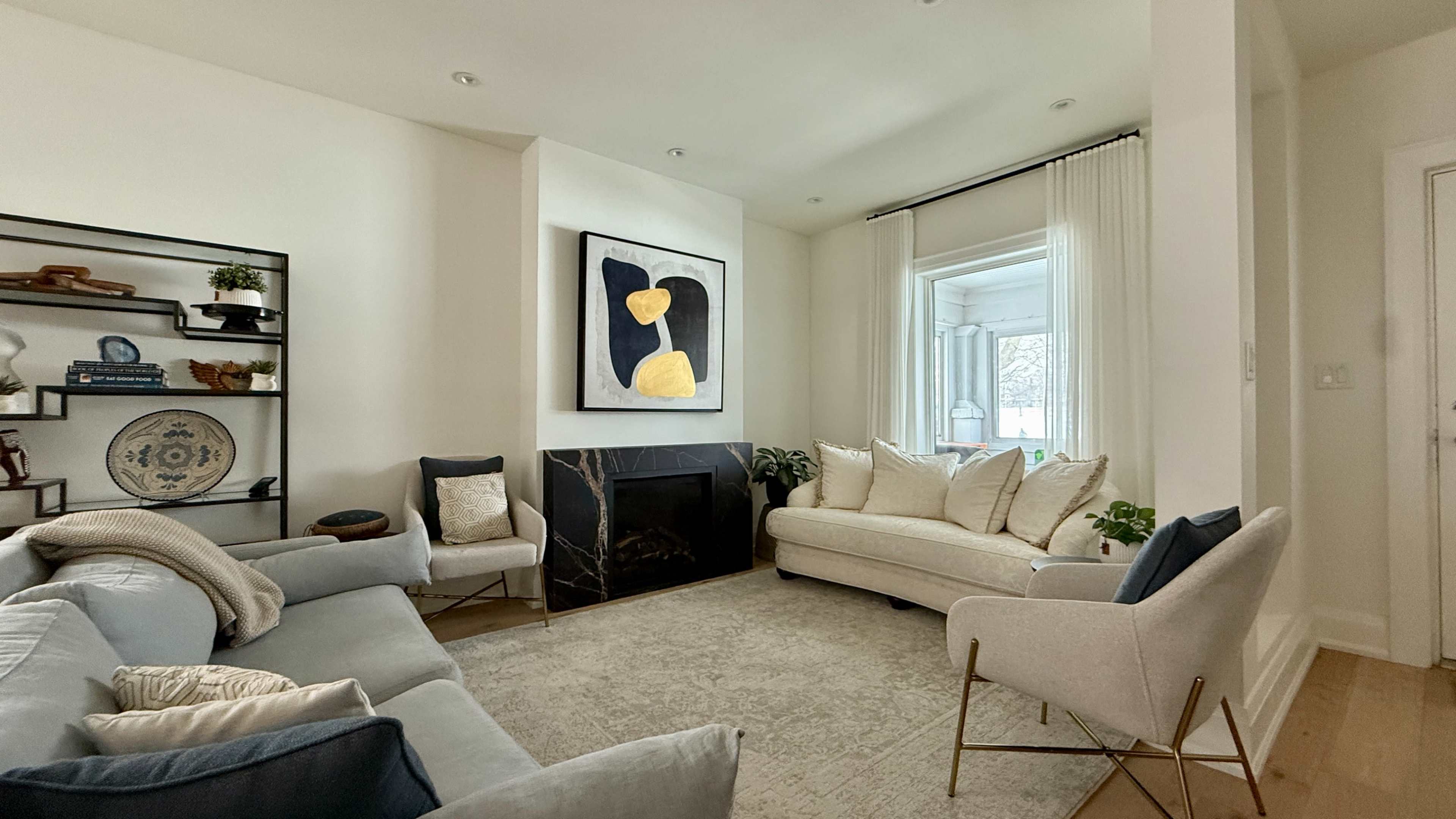 A modern living room features a light color palette with a blue sofa, a cream-colored couch, an area rug, and large windows with sheer curtains.