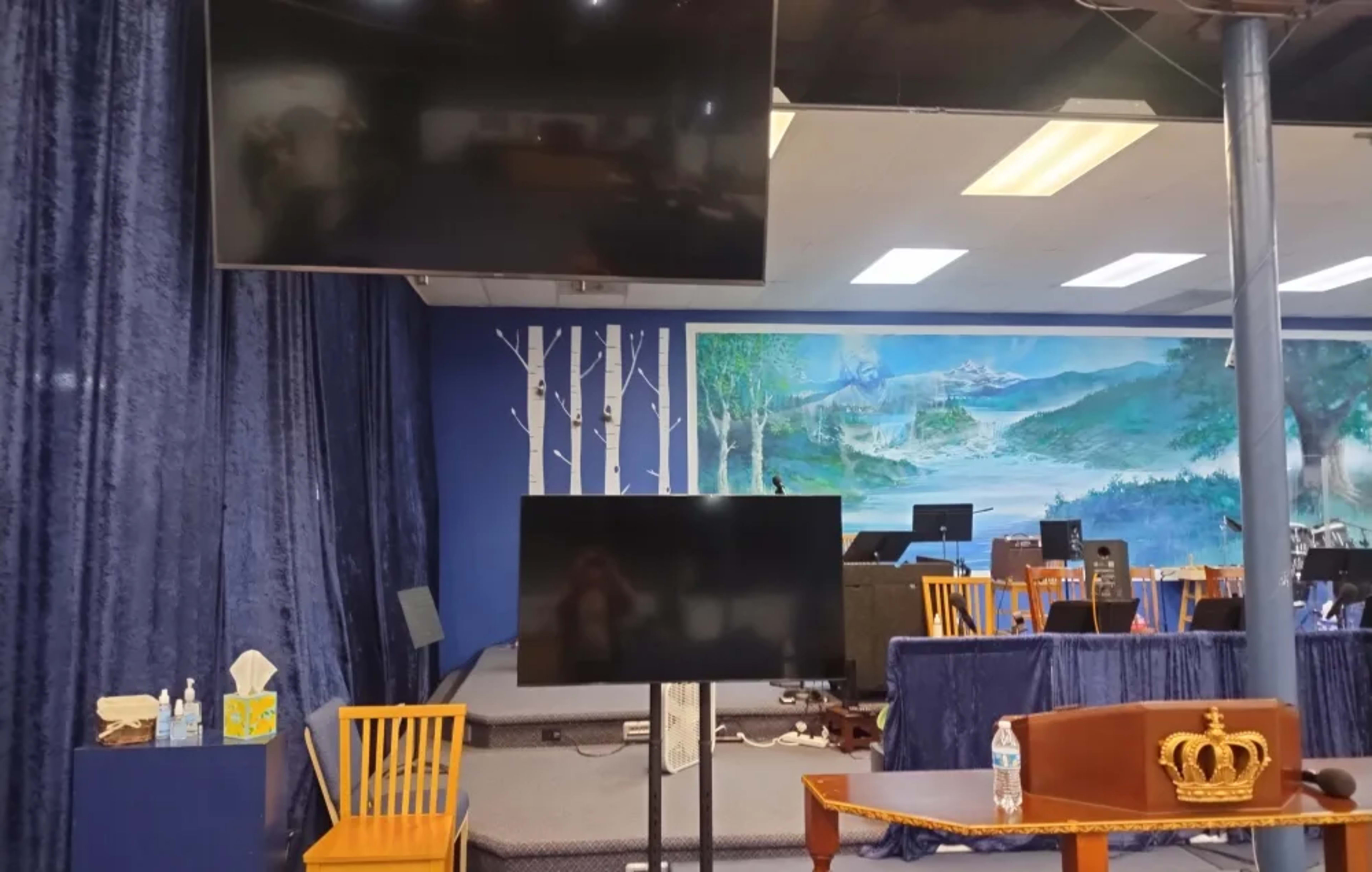 The image shows a stage area with two large screens mounted on the wall, a wooden chair, a bottle of water, and a decorative crown on a table.