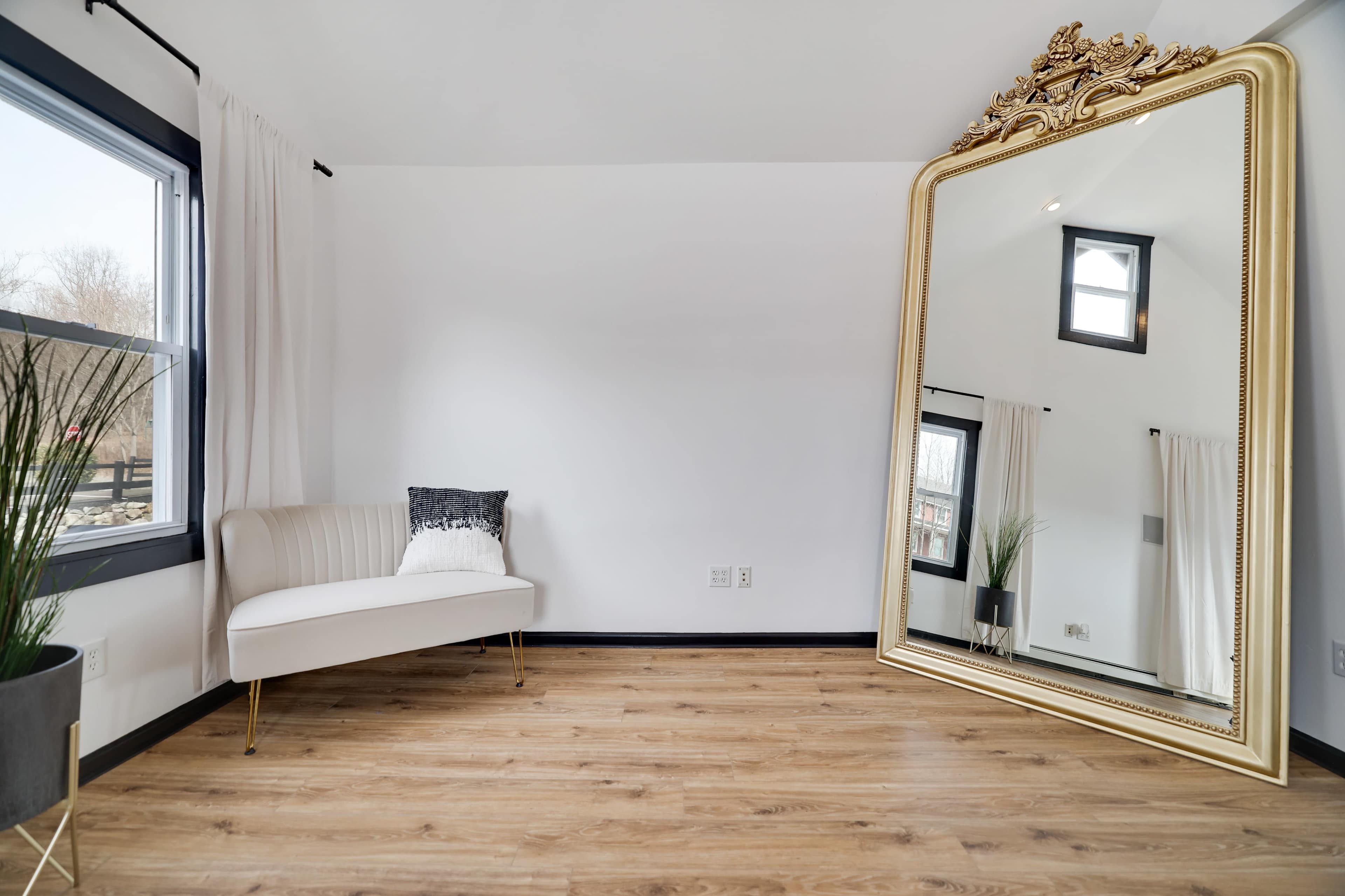 A spacious room features a large ornate mirror, a white couch, and large windows with natural light.
