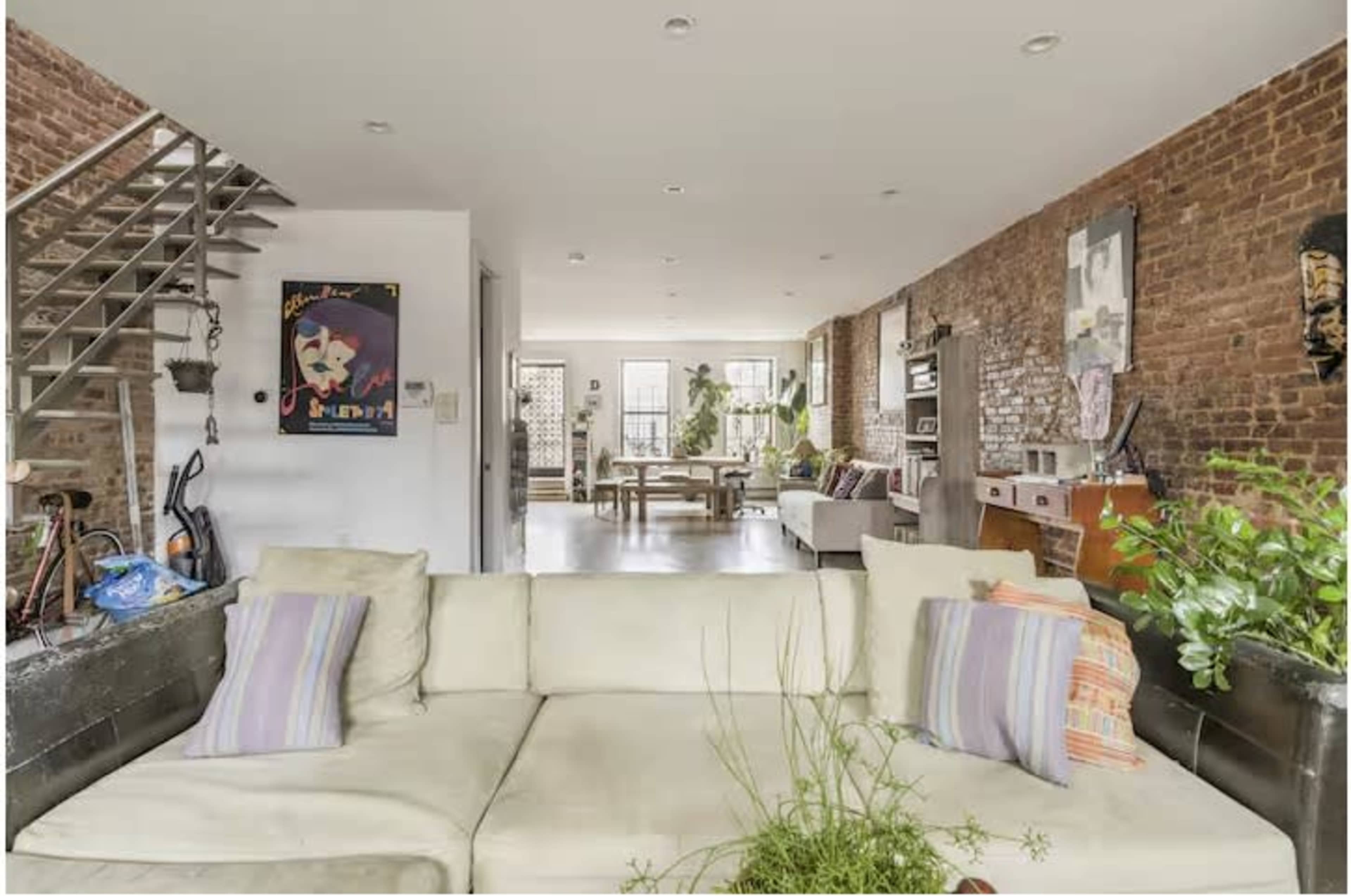 The image shows a modern living space with exposed brick walls, a light-colored sofa, a dining area, and various indoor plants.