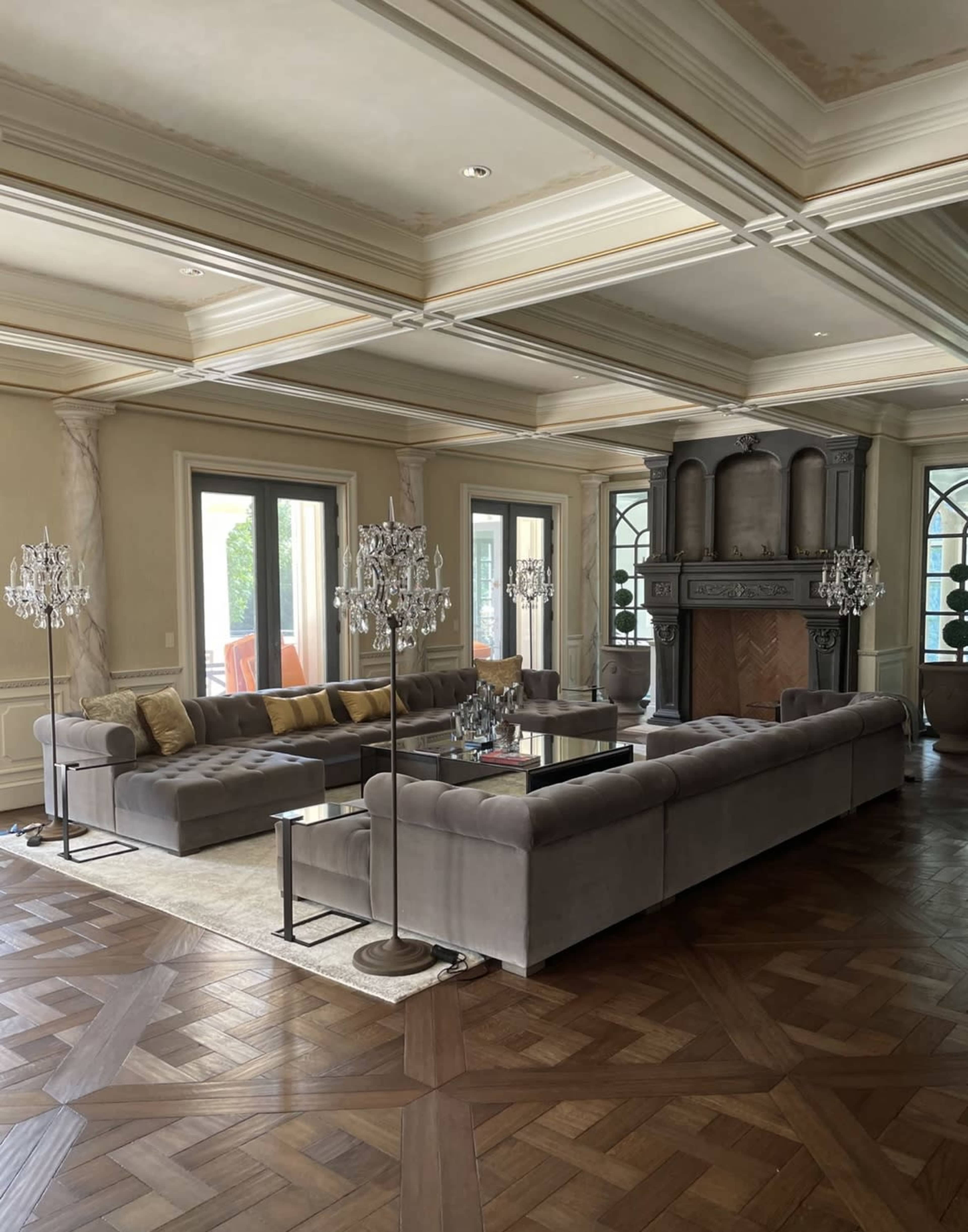 A spacious living room features a large sectional sofa, a central coffee table, and decorative chandeliers beside a fireplace, with large windows providing natural light.