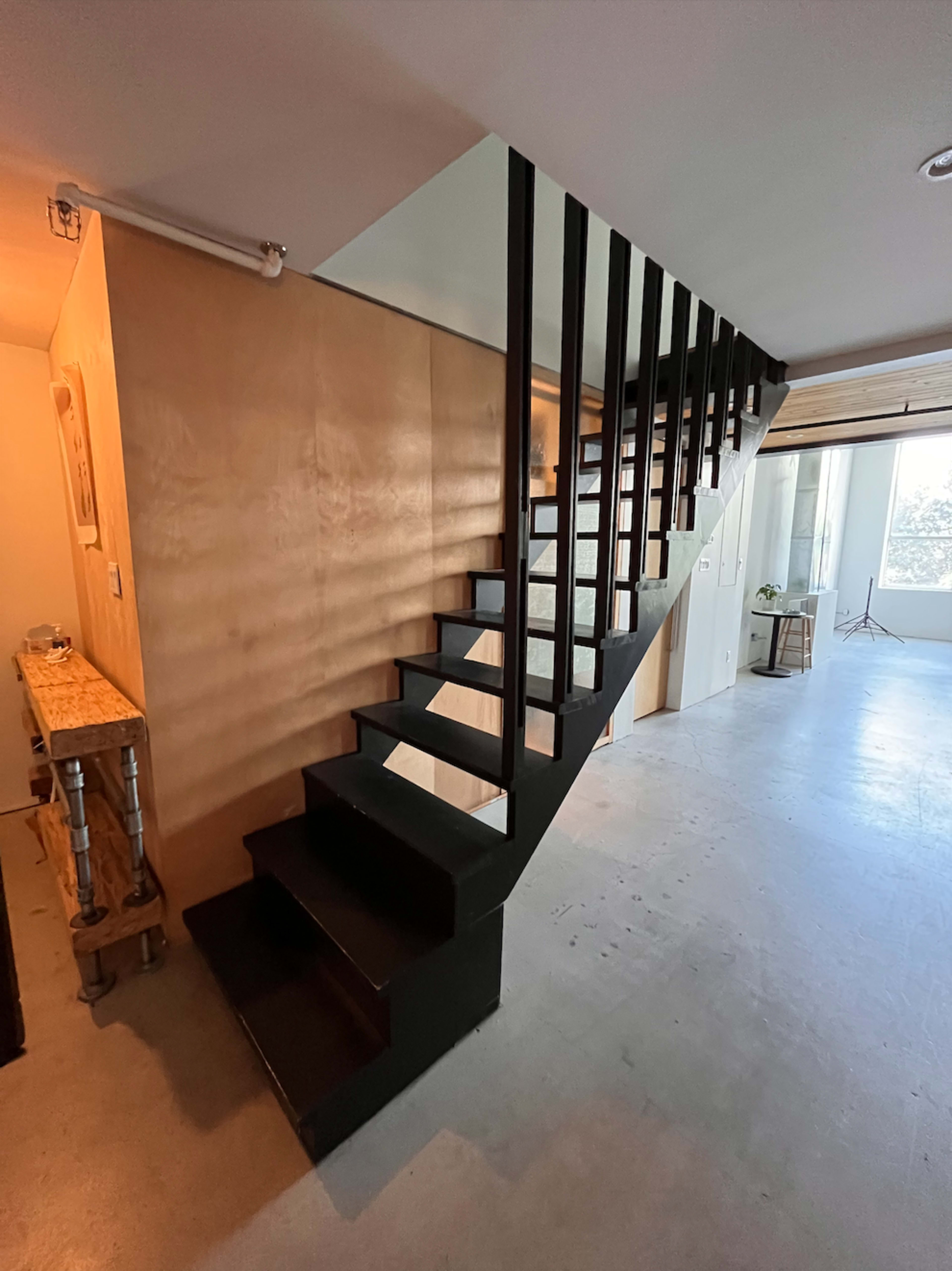 A black staircase ascends against a light wooden wall in a spacious room with large windows.