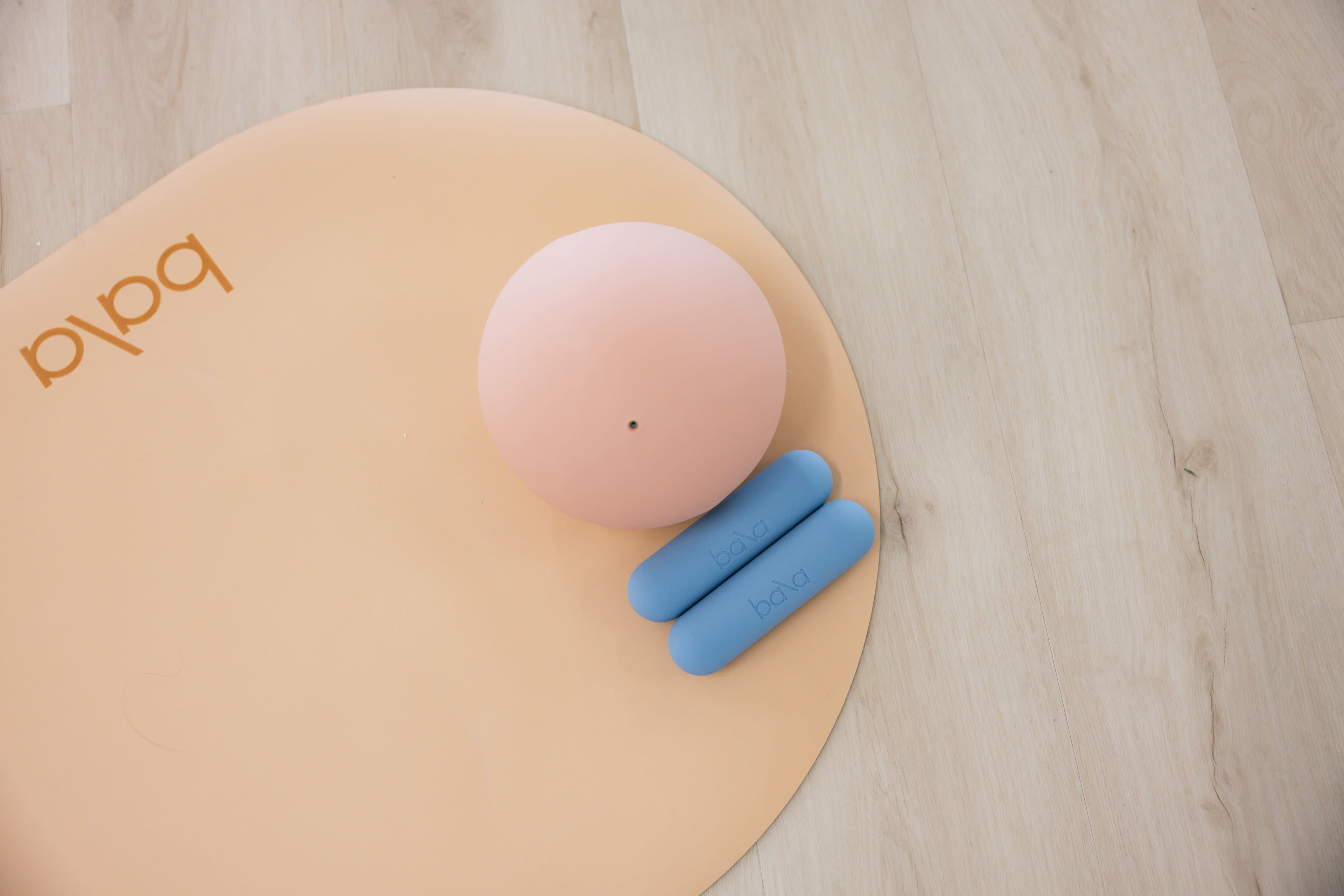 The image shows a peach-colored yoga mat with a pink exercise ball and a pair of blue hand weights placed on it.