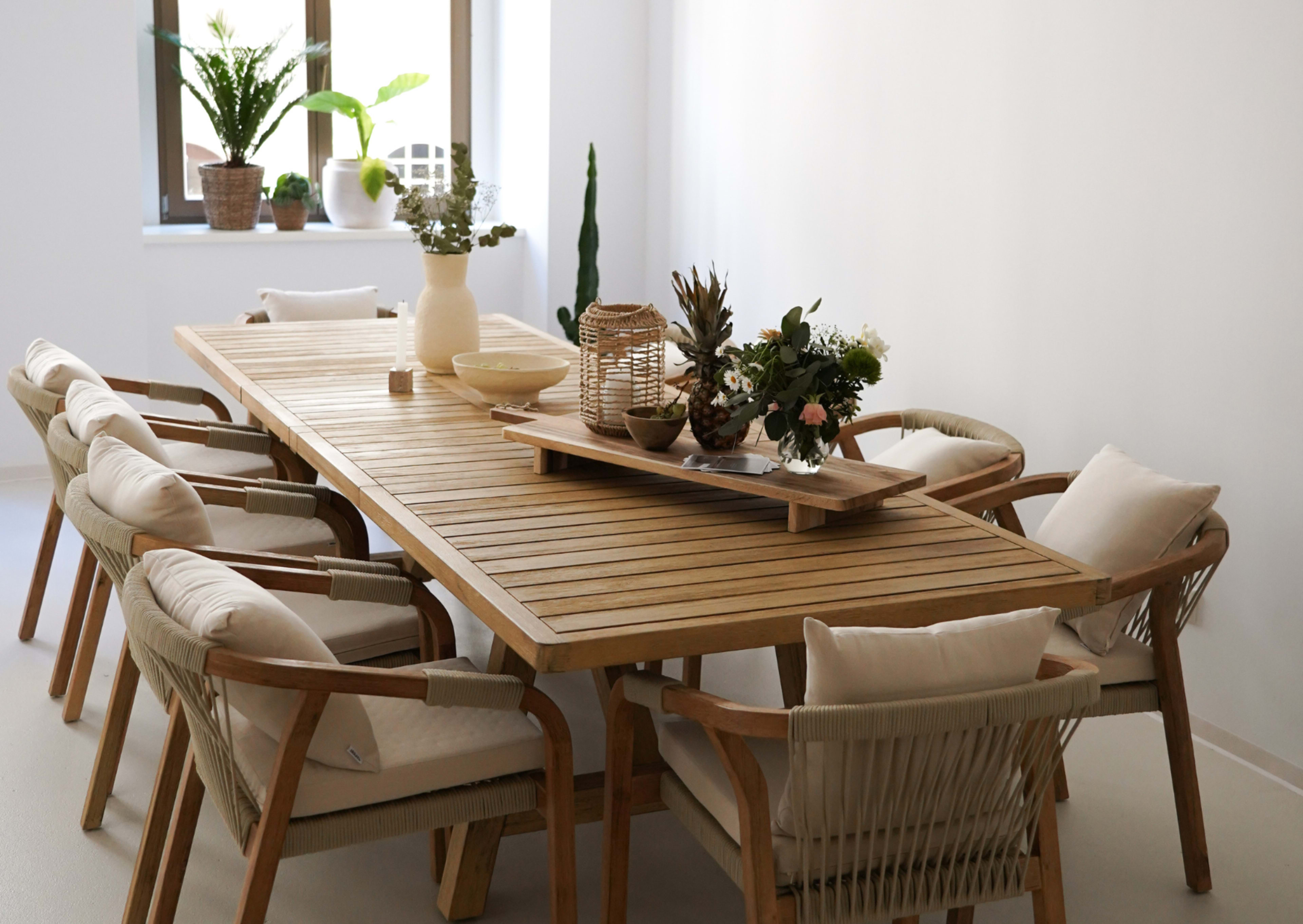 A wooden dining table with eight chairs, decorated with plants and a centerpiece, is set in a bright, minimalistic room.
