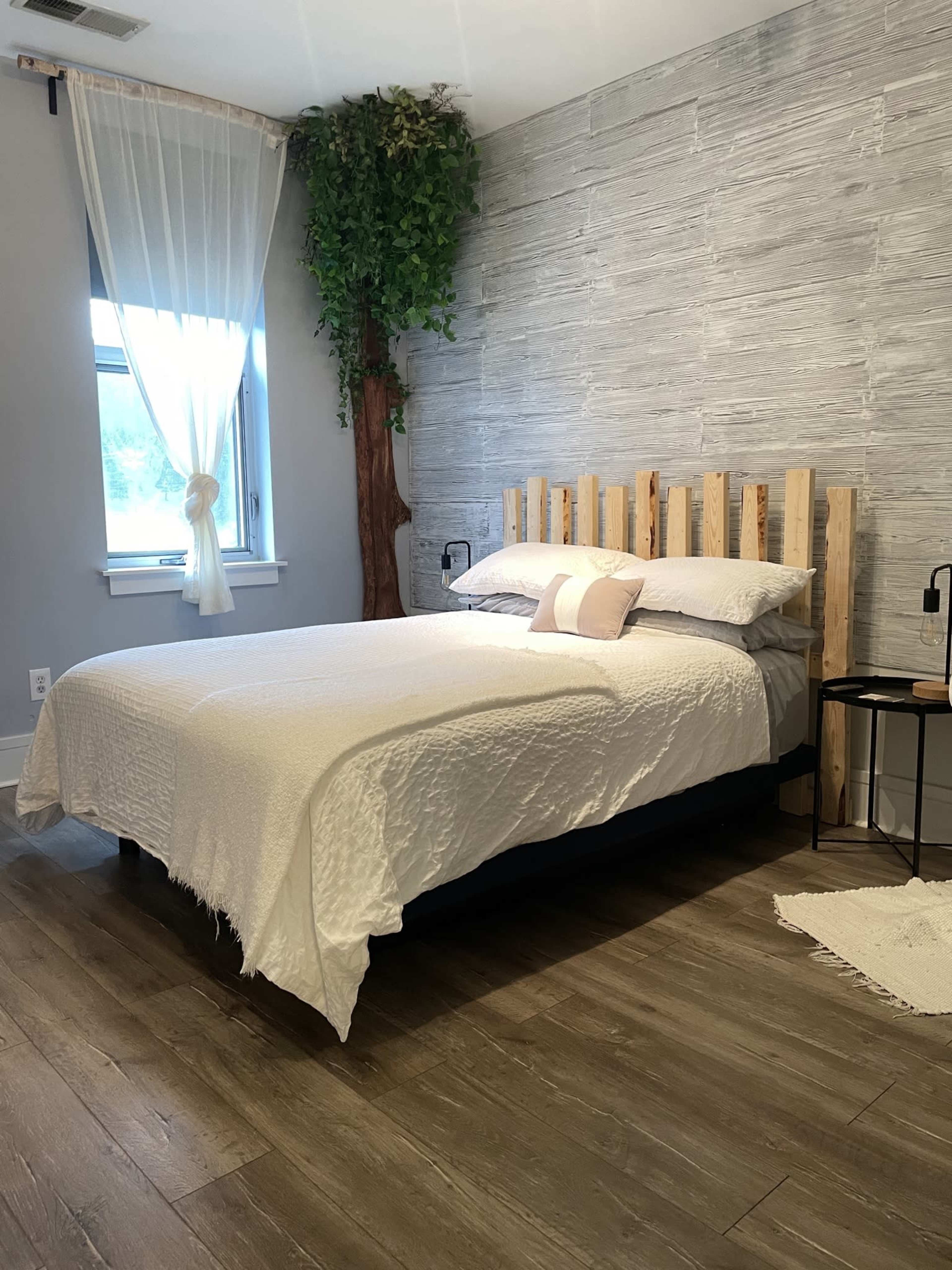 A neatly made bed with a white bedspread is situated in a room featuring a wooden accent wall, large window, and a decorative tree.