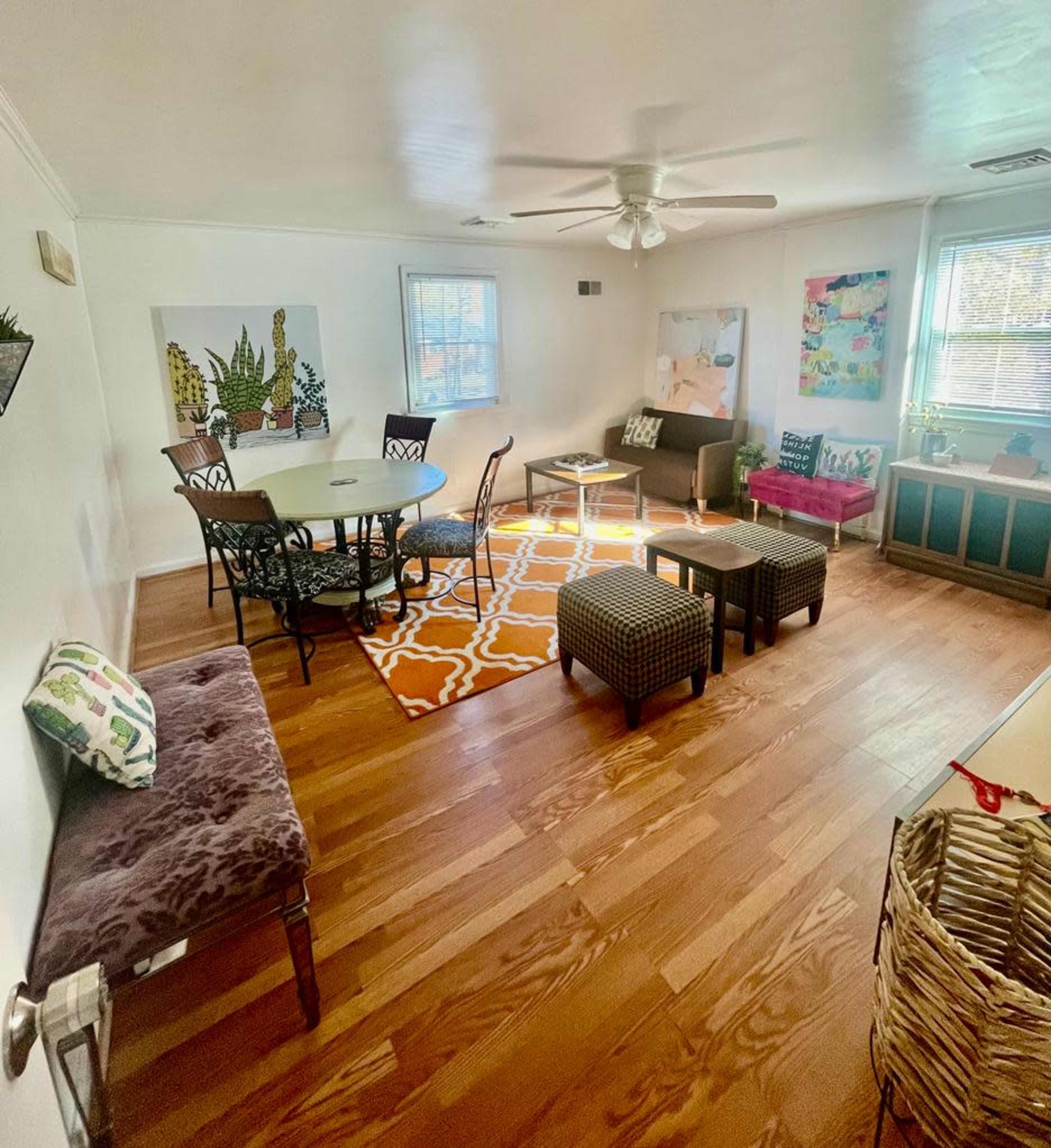 The living room features hardwood floors, a round dining table, multiple seating areas, and colorful artwork on the walls.