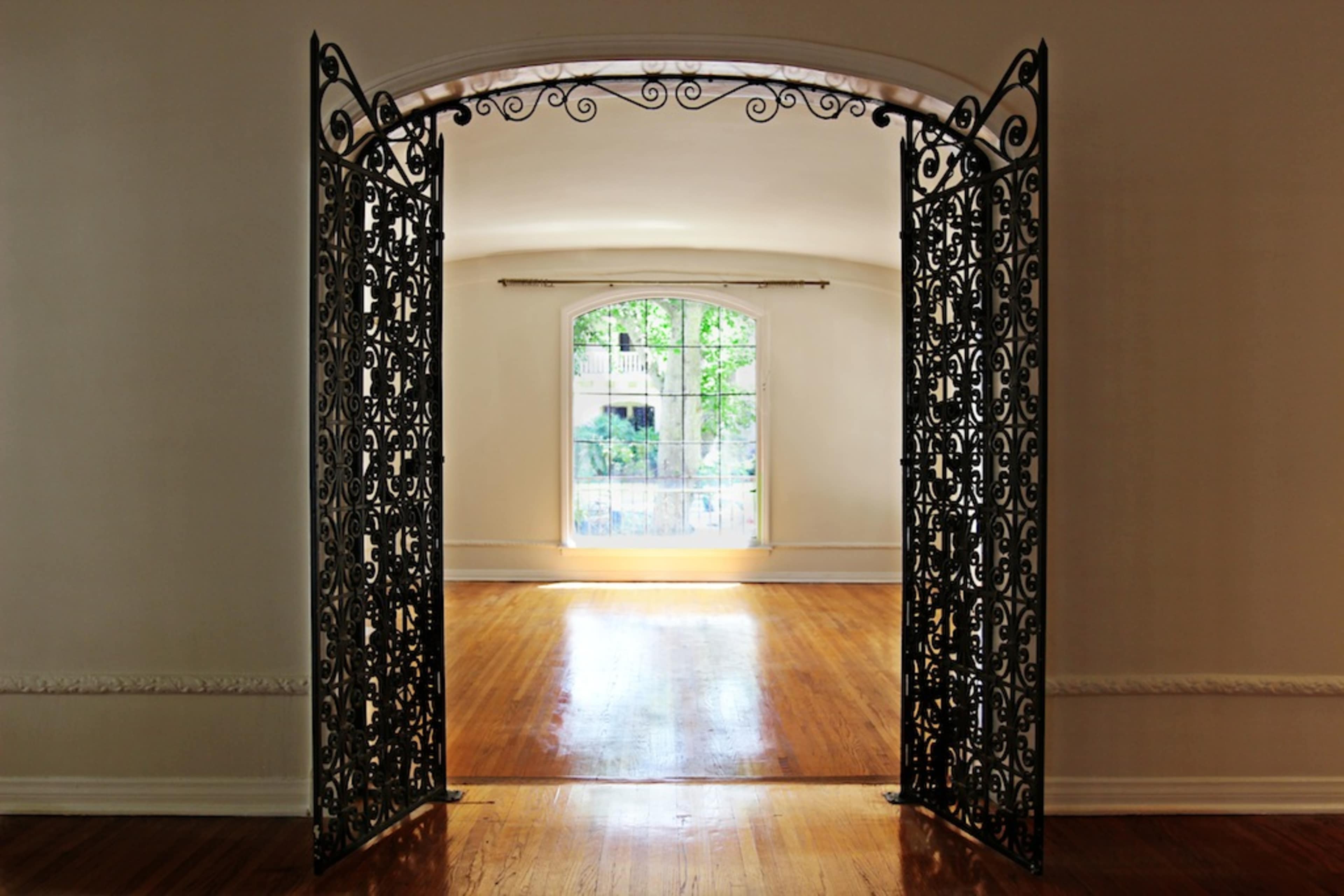 The image shows a pair of ornate wrought iron gates opening into a brightly lit room with a large window.