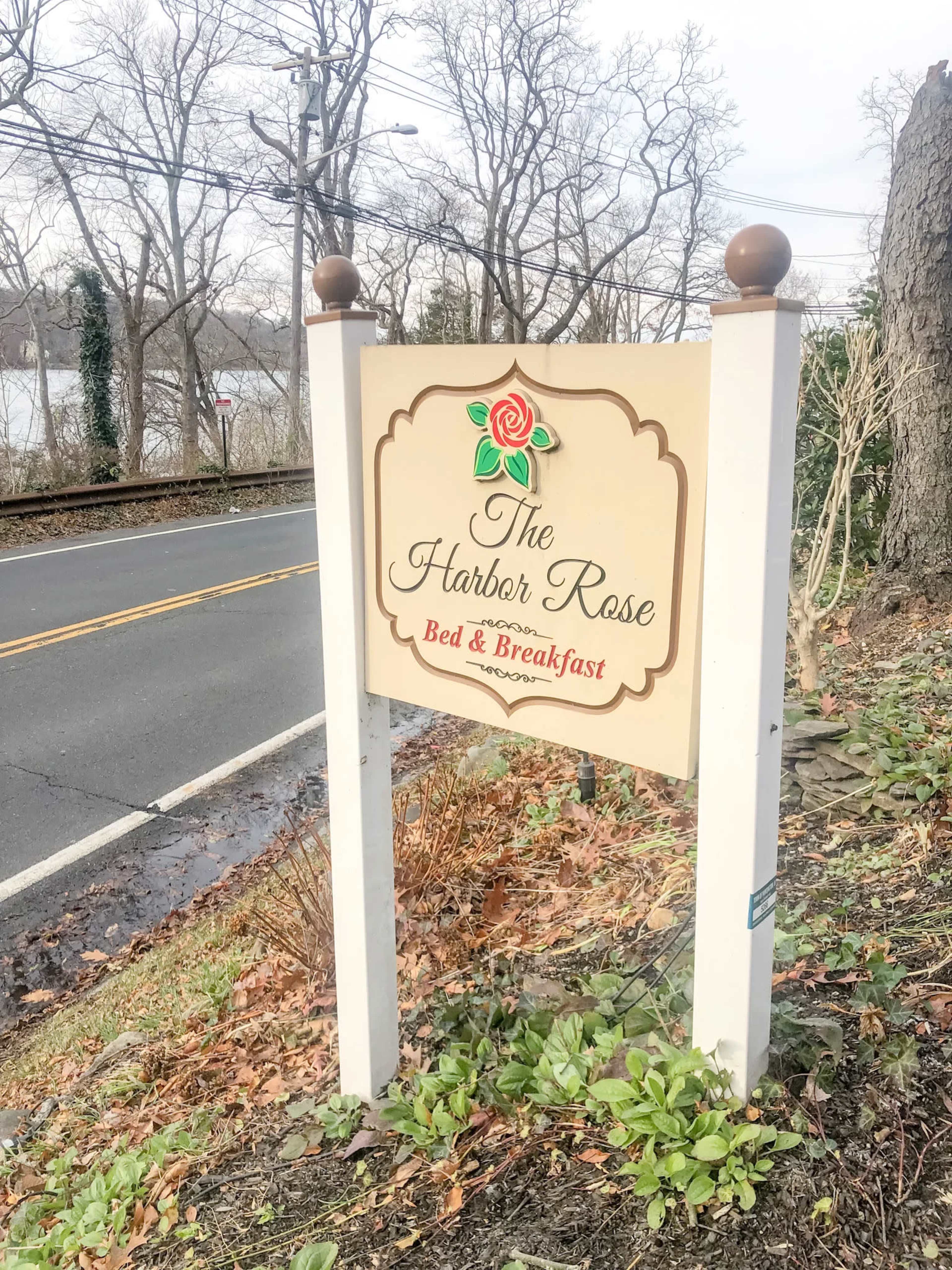 A wooden sign for "The Harbor Rose Bed & Breakfast" stands by the roadside, with trees and a body of water visible in the background.