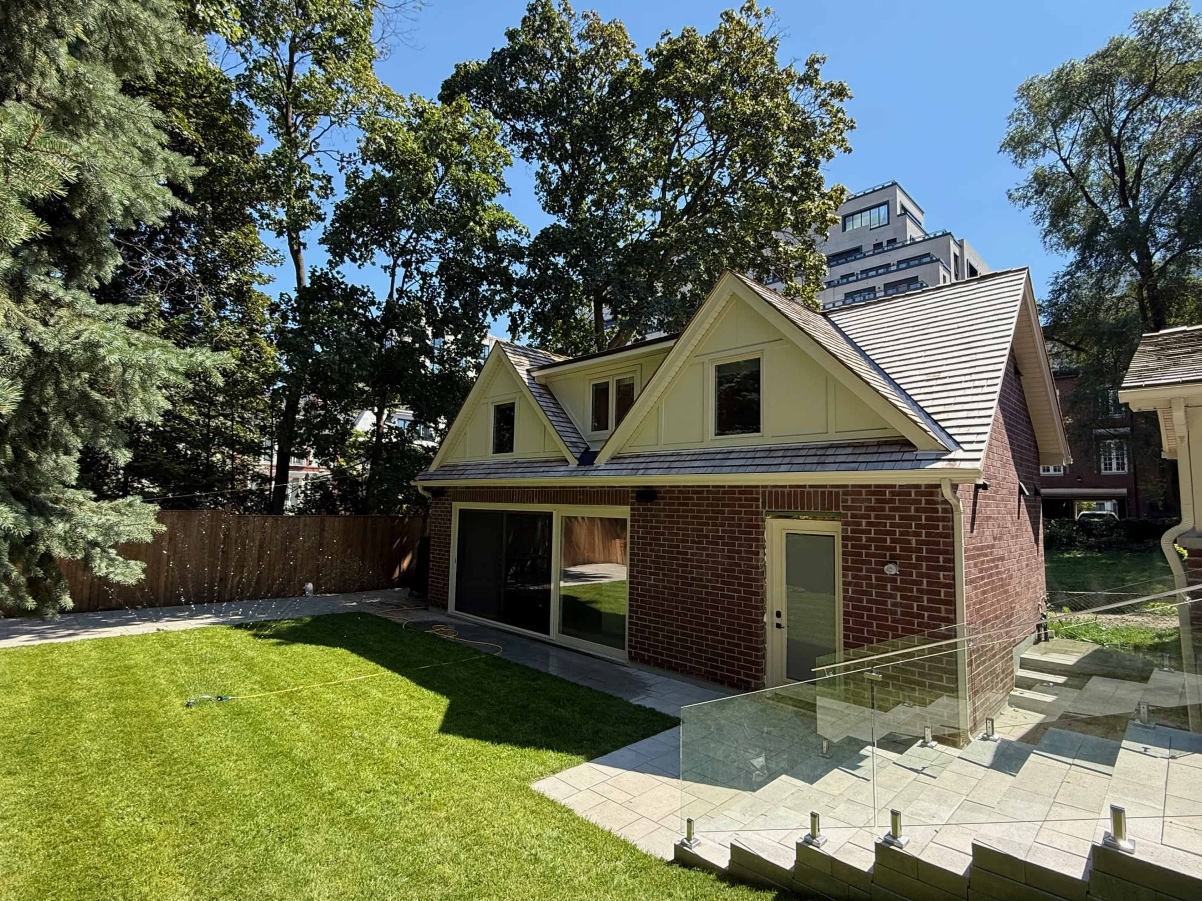 A two-story house with a brick exterior and a sloped roof is situated in a lush backyard surrounded by trees and adjacent to a modern building.