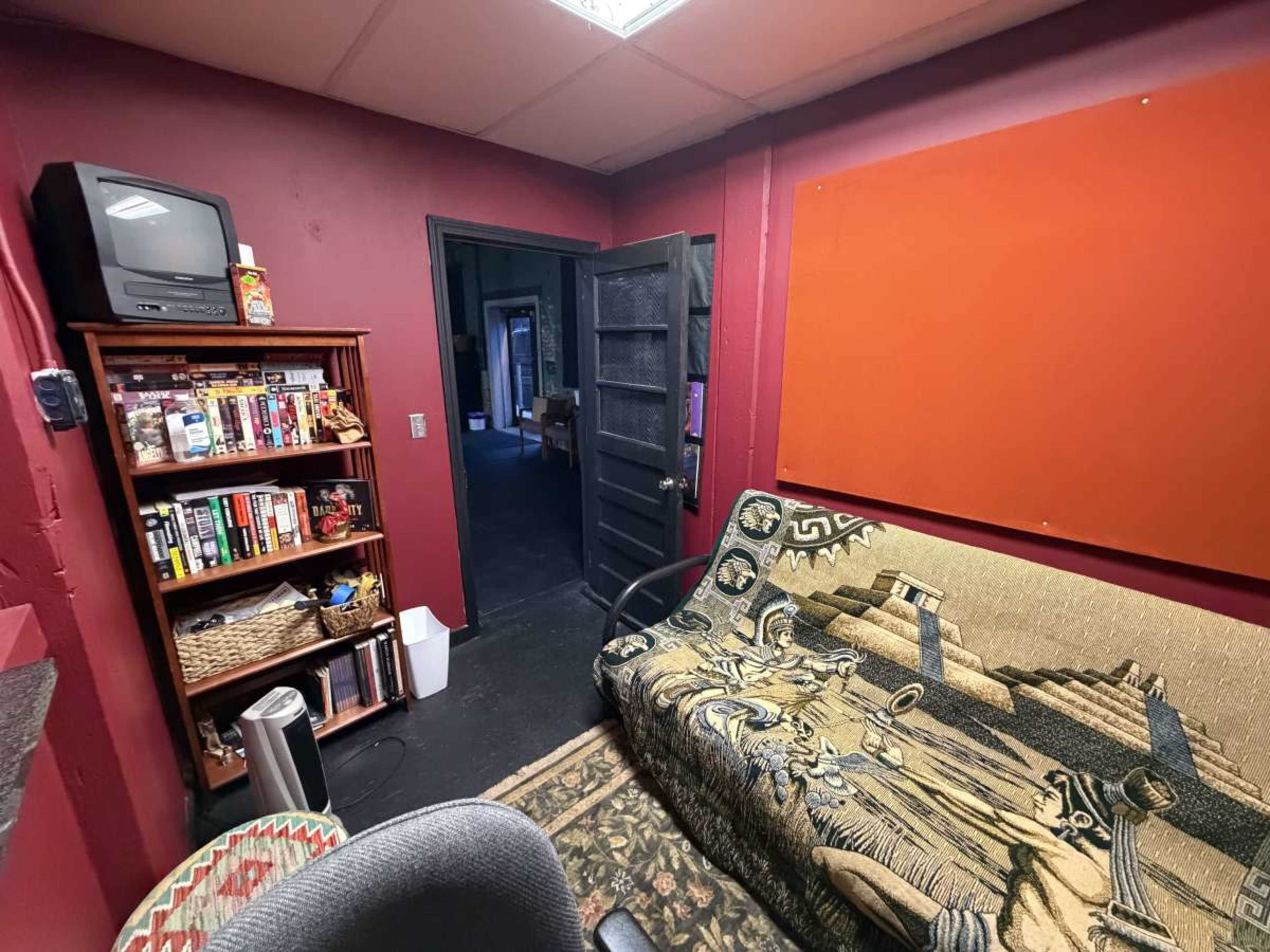The image shows a small dimly lit room with burgundy walls, featuring a bookcase filled with books, a vintage television, a patterned couch, and a door leading to a darker hallway.