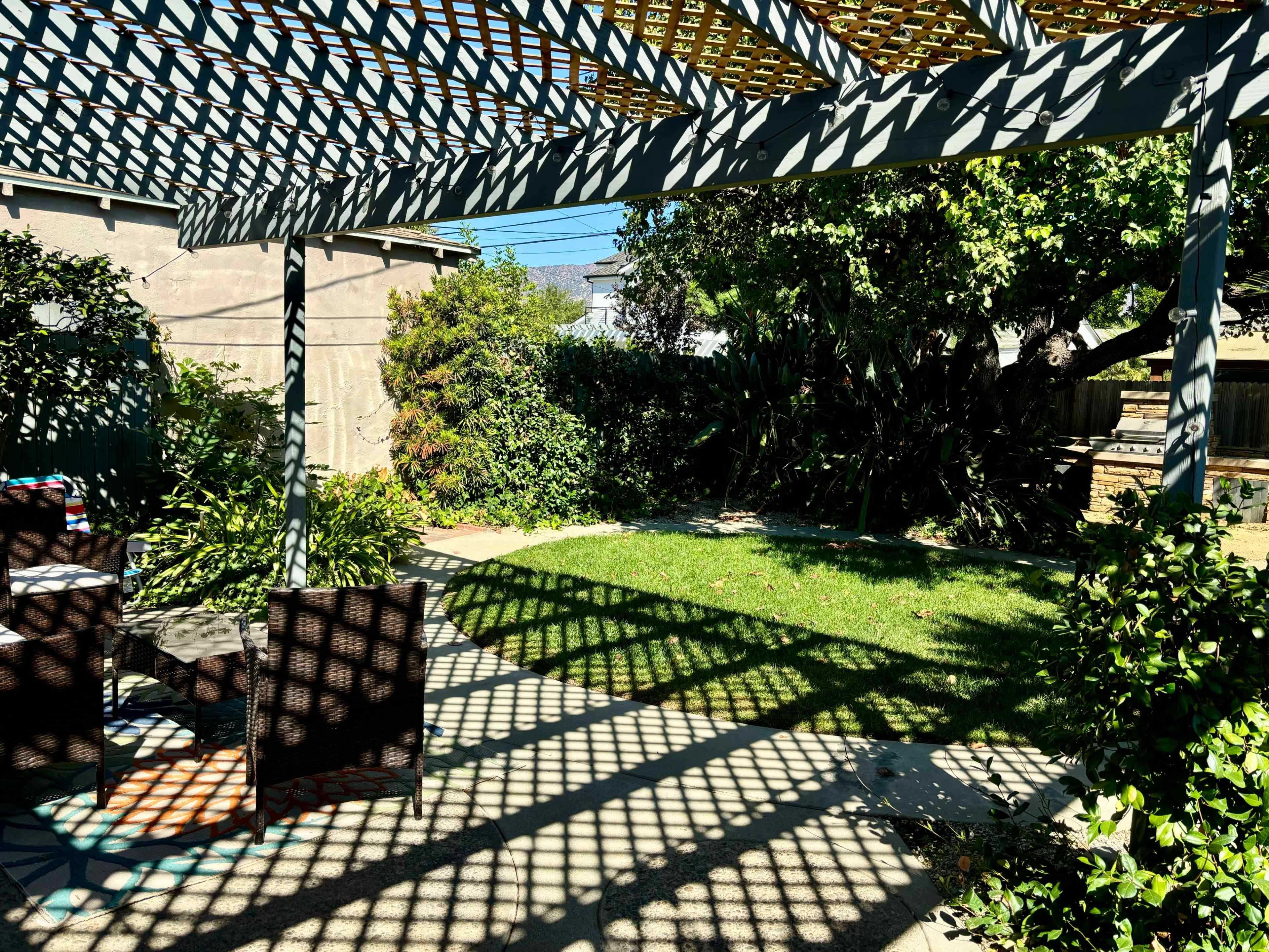 A backyard features a shaded patio area with woven overhead structures and neatly trimmed grass surrounded by greenery.
