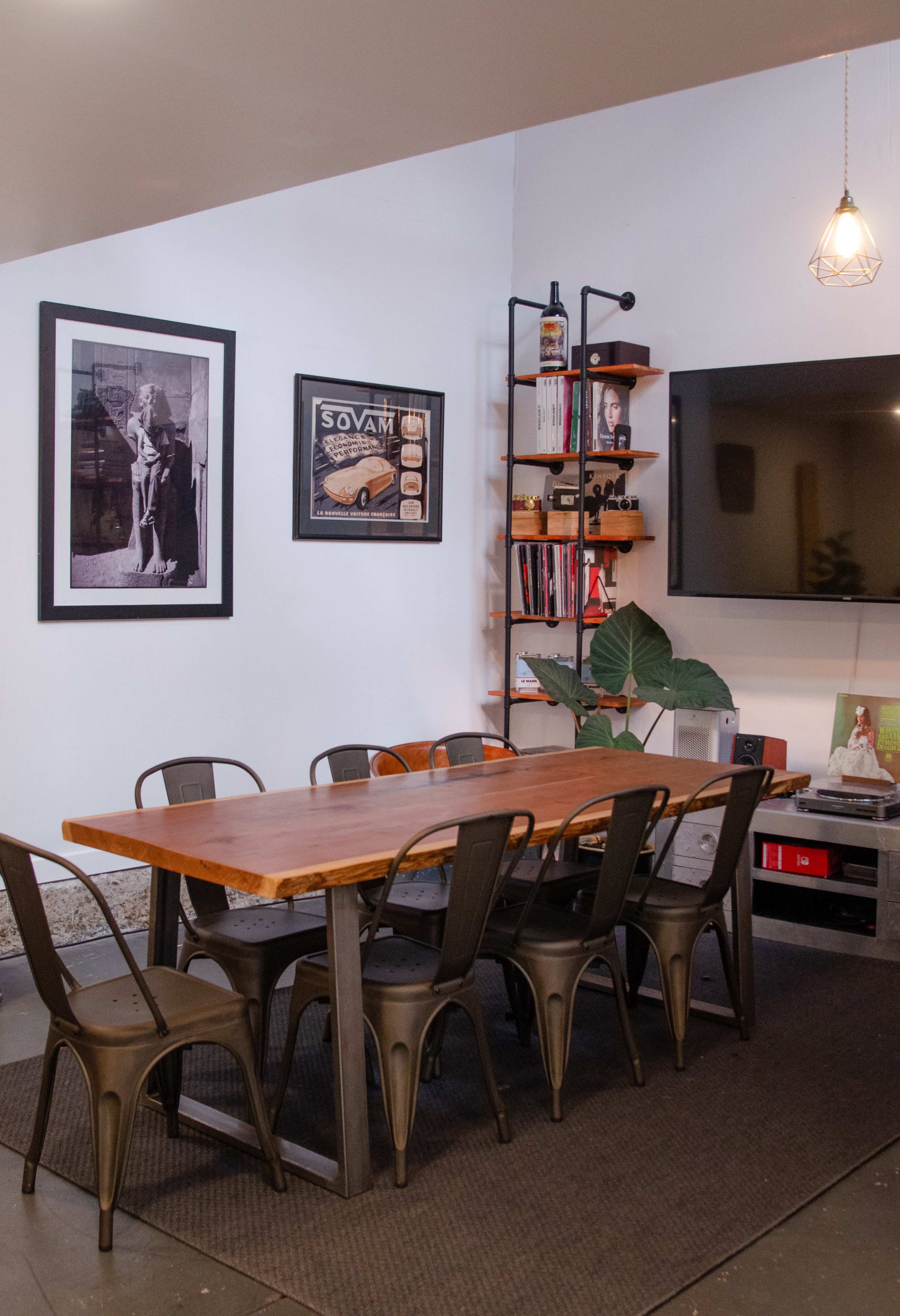 A long wooden dining table with metal chairs is positioned in a room featuring artwork on the walls and a shelf with books and decorative items.