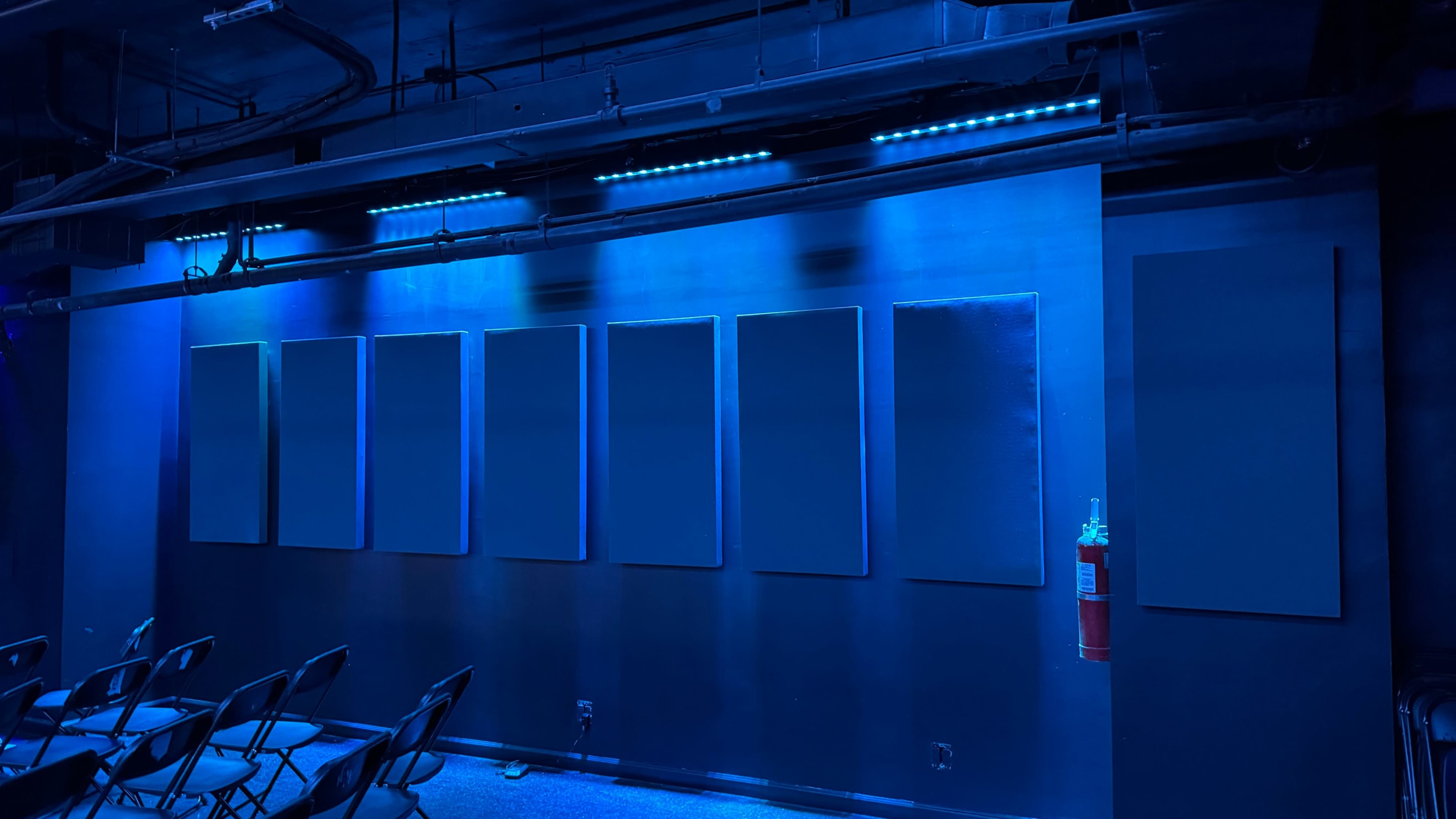 The image shows a dimly lit room with a blue-toned wall featuring seven blank canvases and a fire extinguisher mounted to the side.