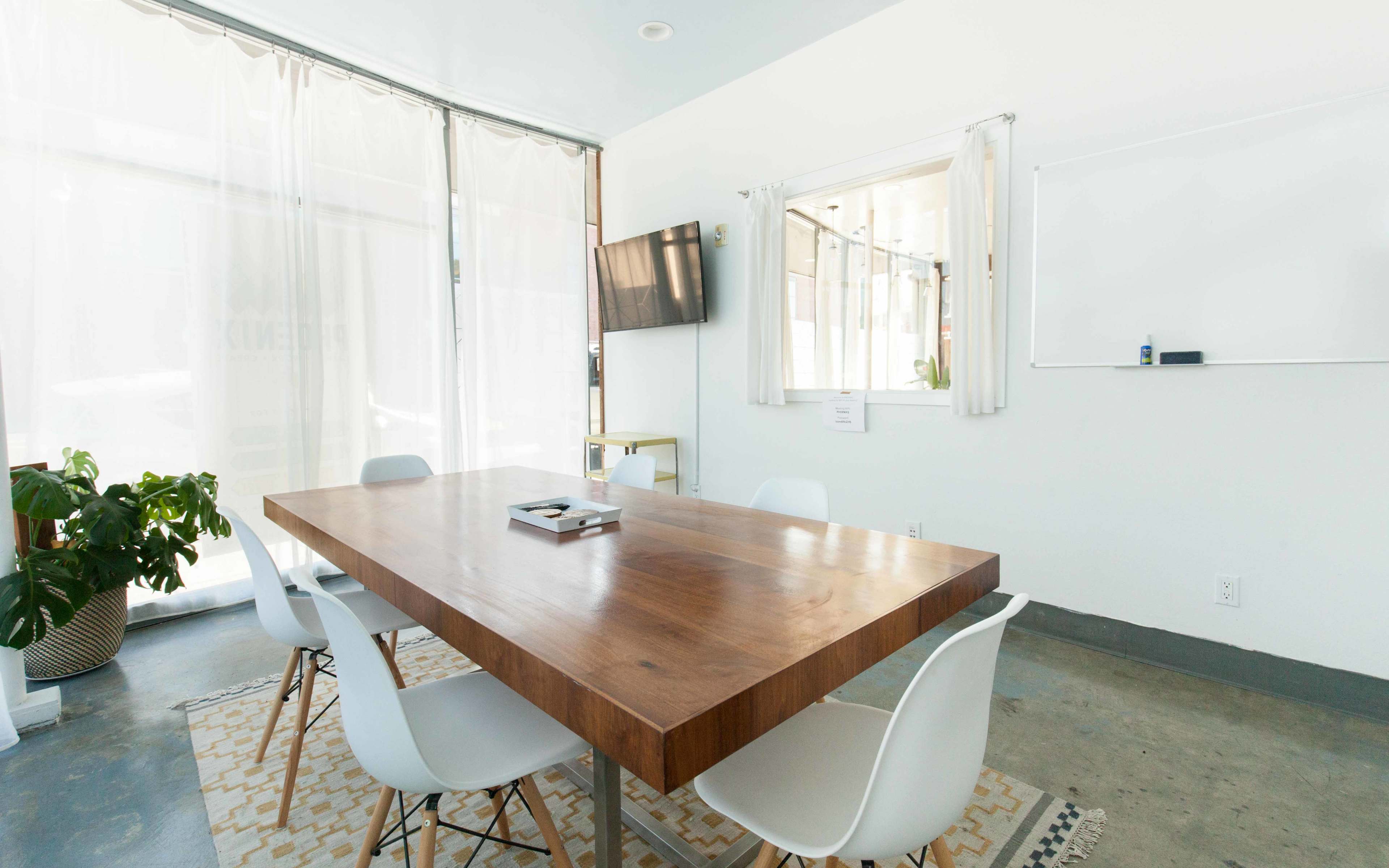 A conference room features a large wooden table surrounded by white chairs, with a television mounted on the wall and natural light filtering through sheer curtains.
