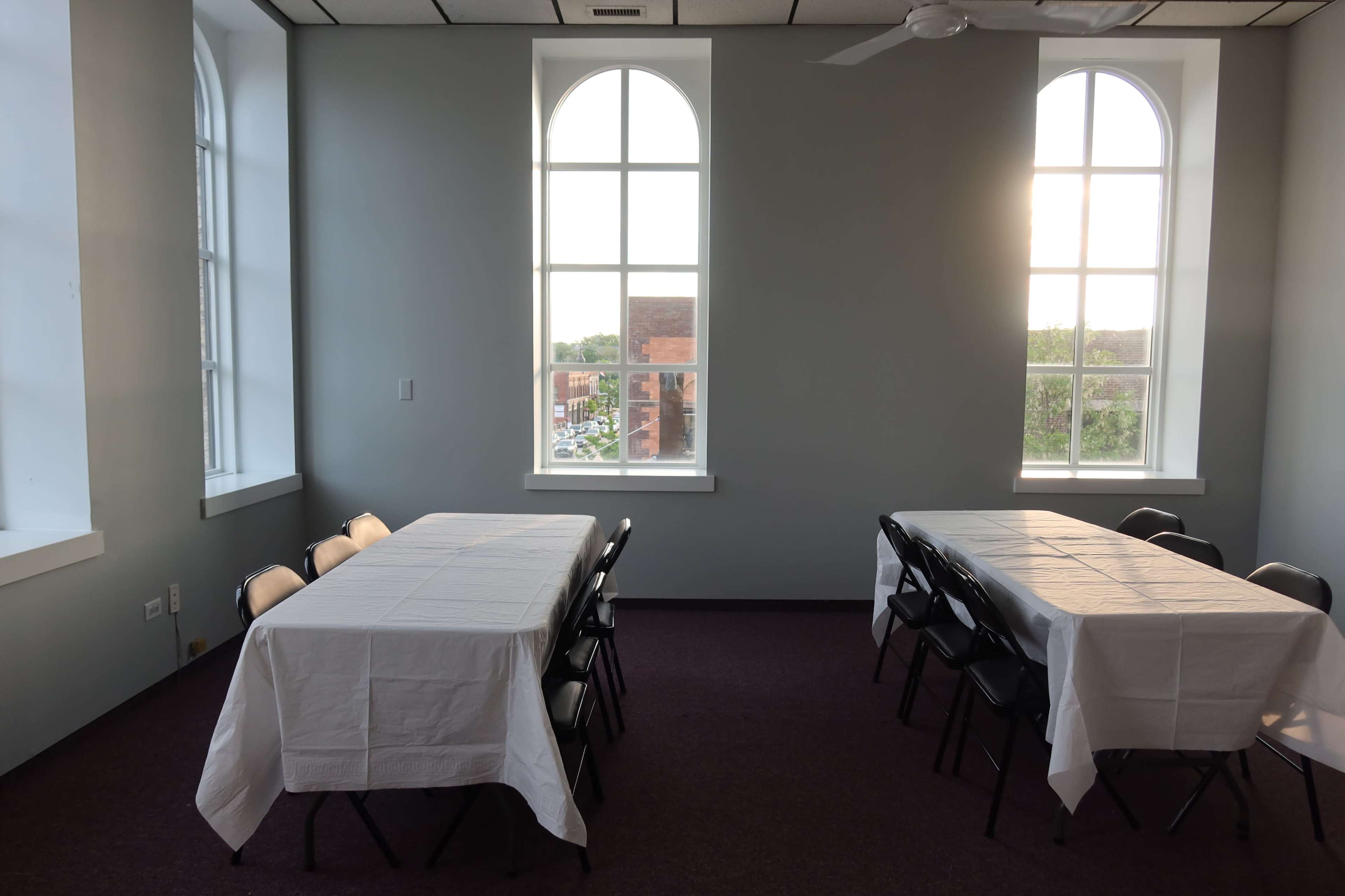 A room with two tables covered in white tablecloths, arranged for a gathering, and large arched windows letting in natural light.
