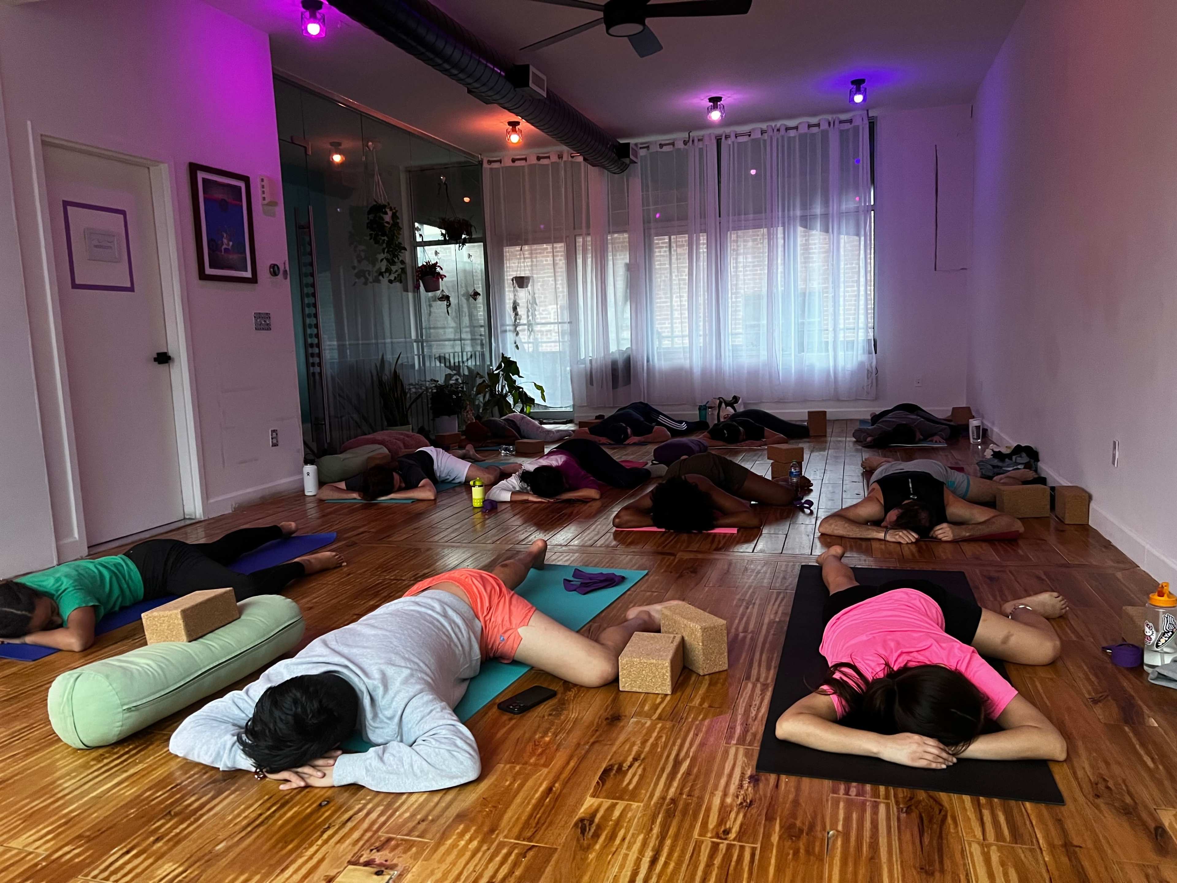 A group of people are practicing yoga in a softly lit studio, lying on mats with props around them.