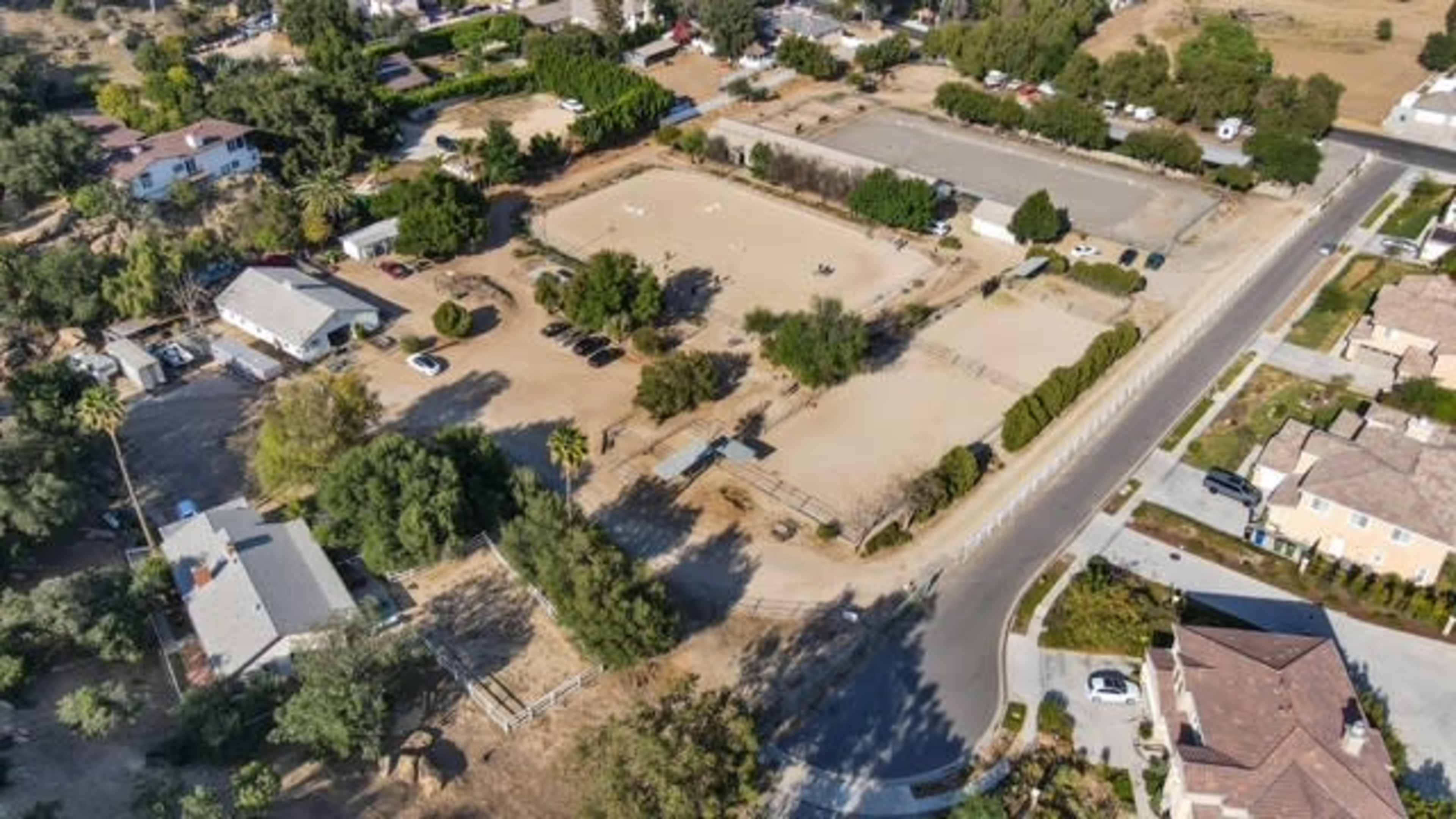 The image shows an aerial view of a rural property with an equestrian arena, surrounded by houses and trees, along with nearby roads.
