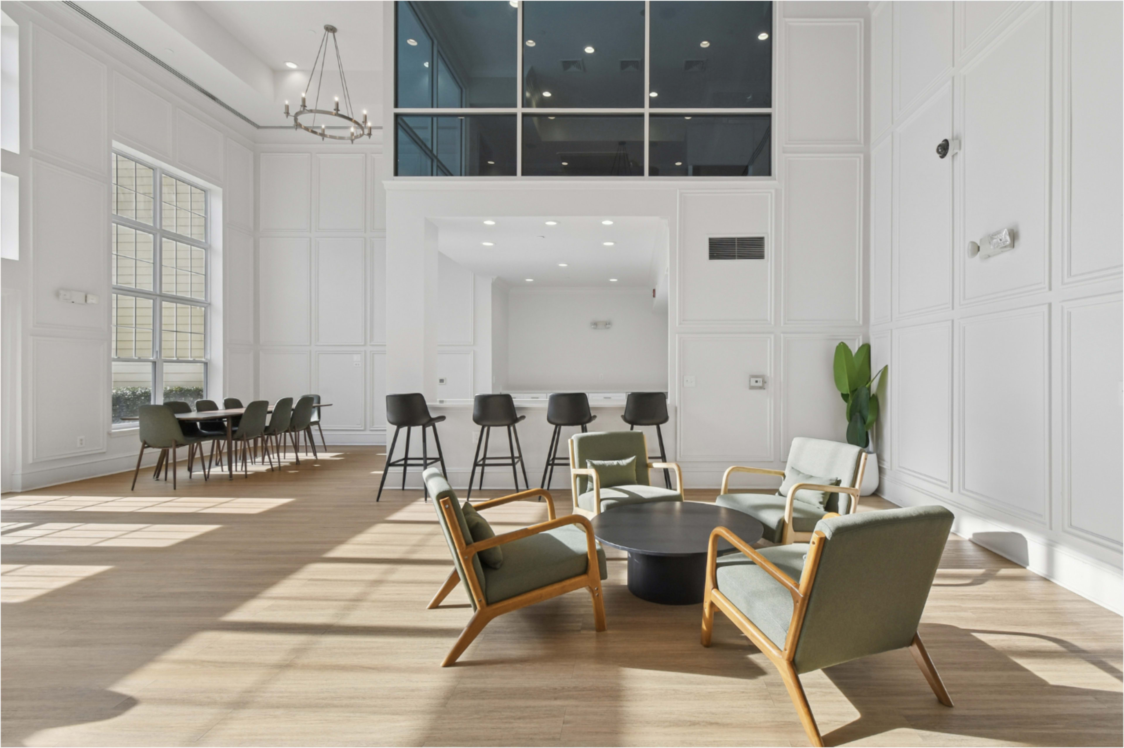 The image shows a modern, brightly lit lounge area featuring a round table surrounded by four armchairs and a row of barstools against the wall.