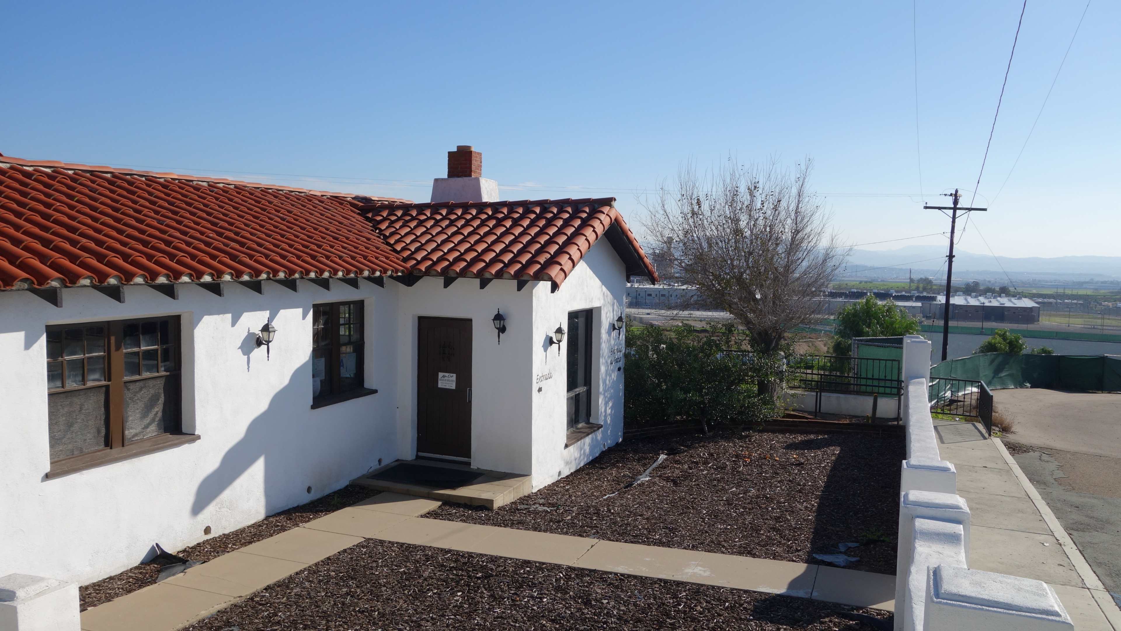 A white stucco house with a red tiled roof is positioned on a sloped lot, with a view of a distant industrial area.