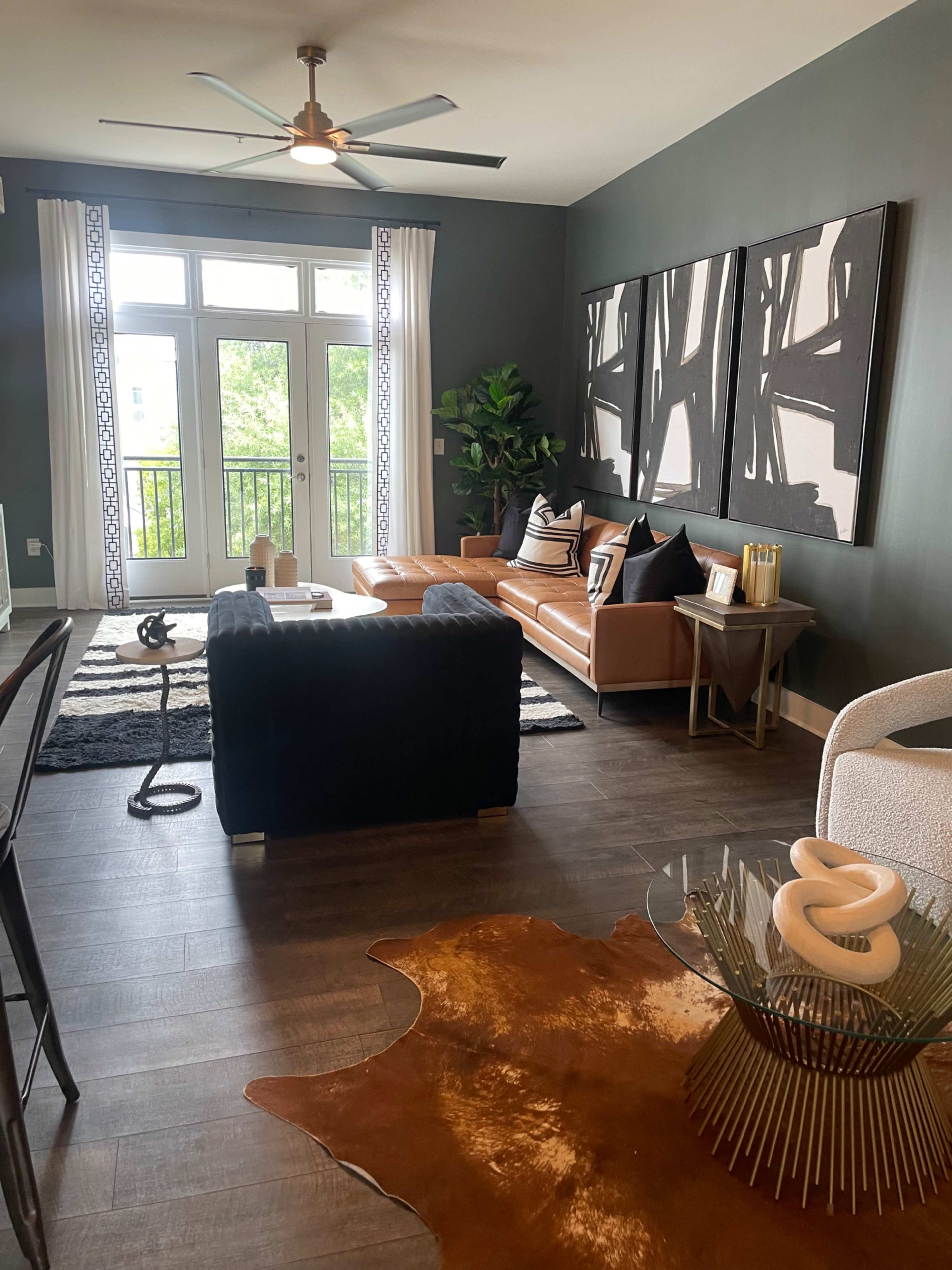 The image shows a modern living room with a leather sofa, a black velvet chair, and a decorative glass coffee table on a cowhide rug, along with large windows that allow natural light to enter the space.