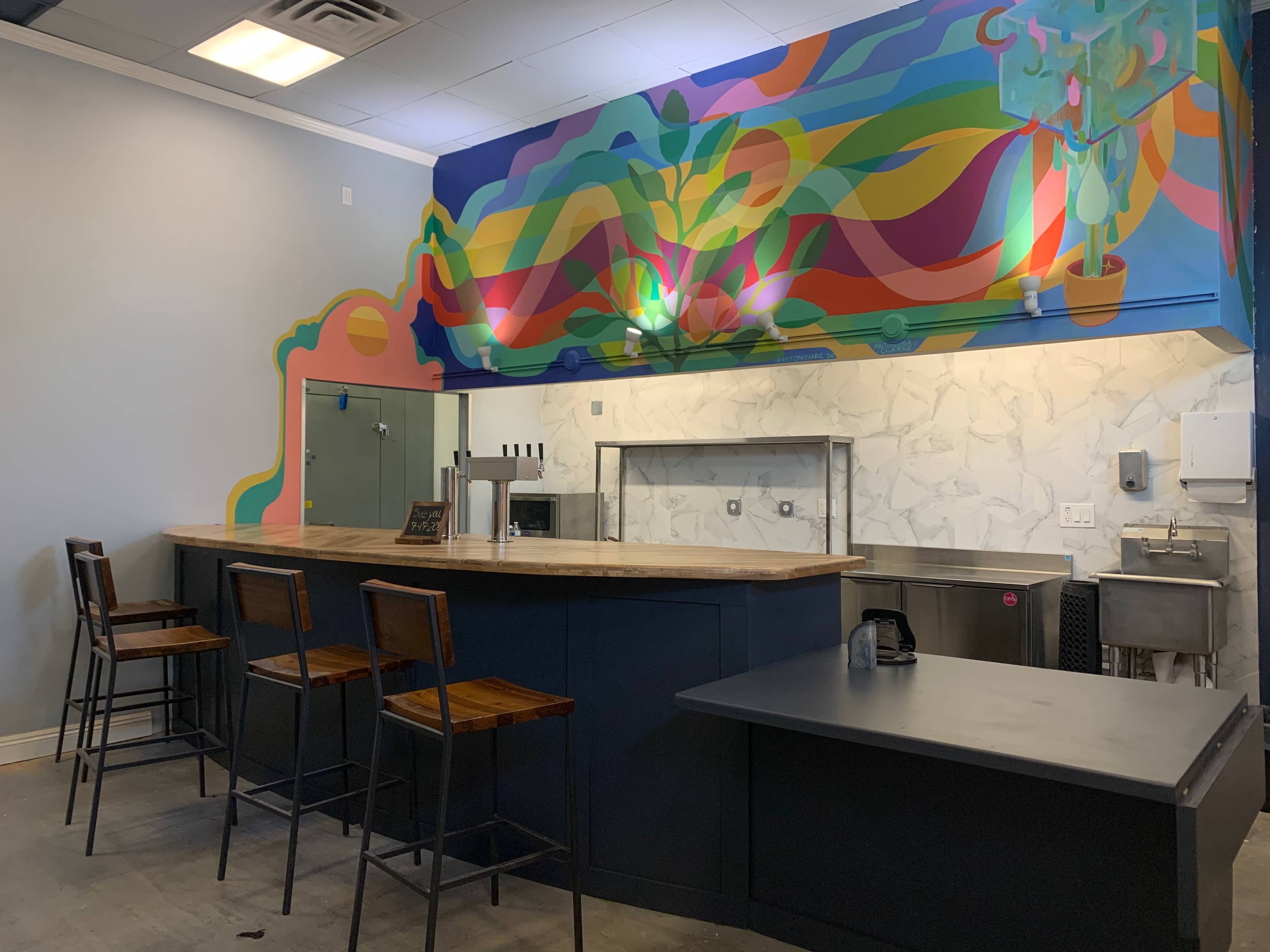 A modern bar area with a colorful mural on the wall, featuring wooden stools and a light-colored countertop.