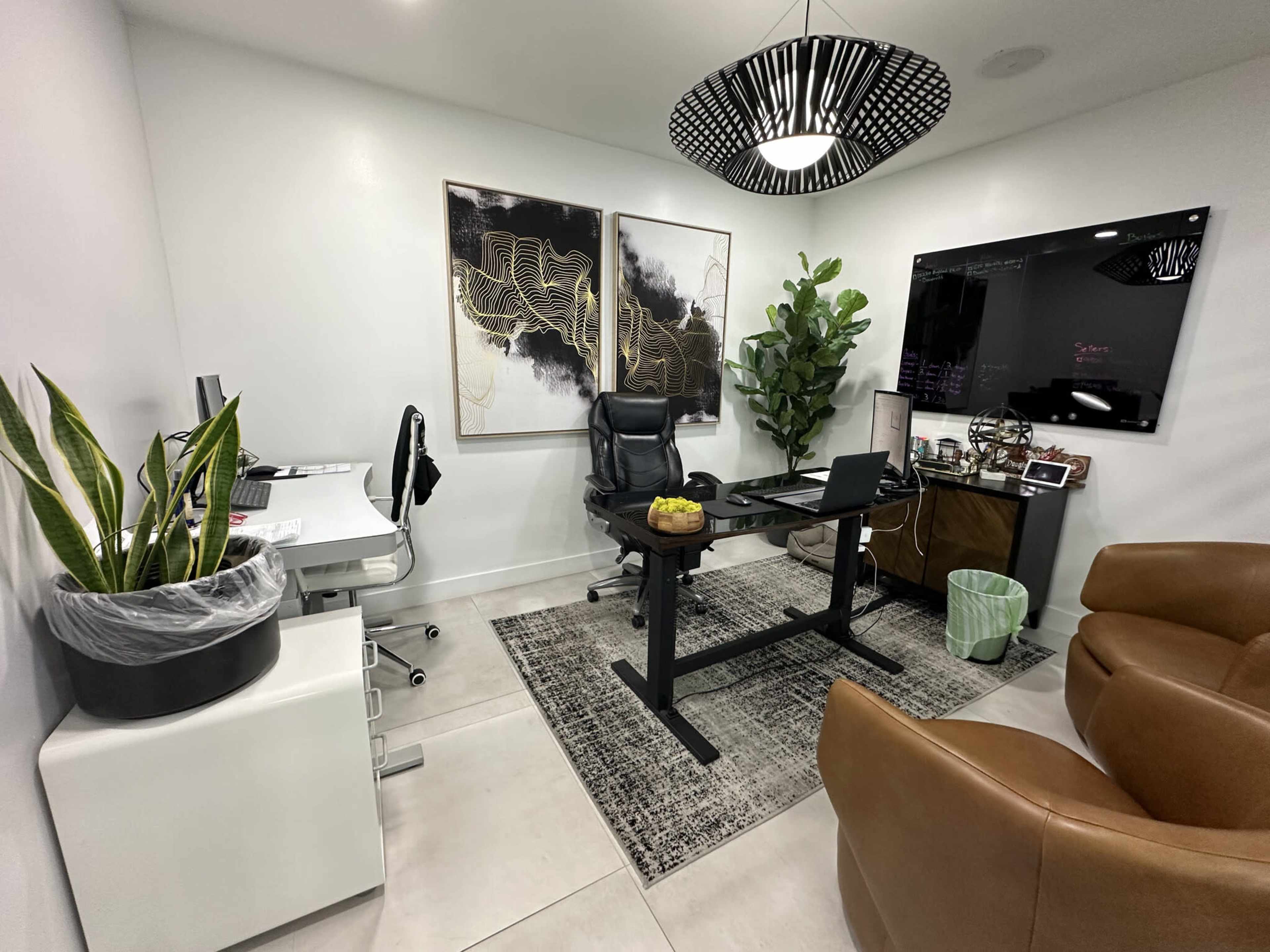 The image shows a modern office space featuring a desk with a chair, a large wall-mounted screen, two abstract paintings, a potted plant, and a brown leather chair.