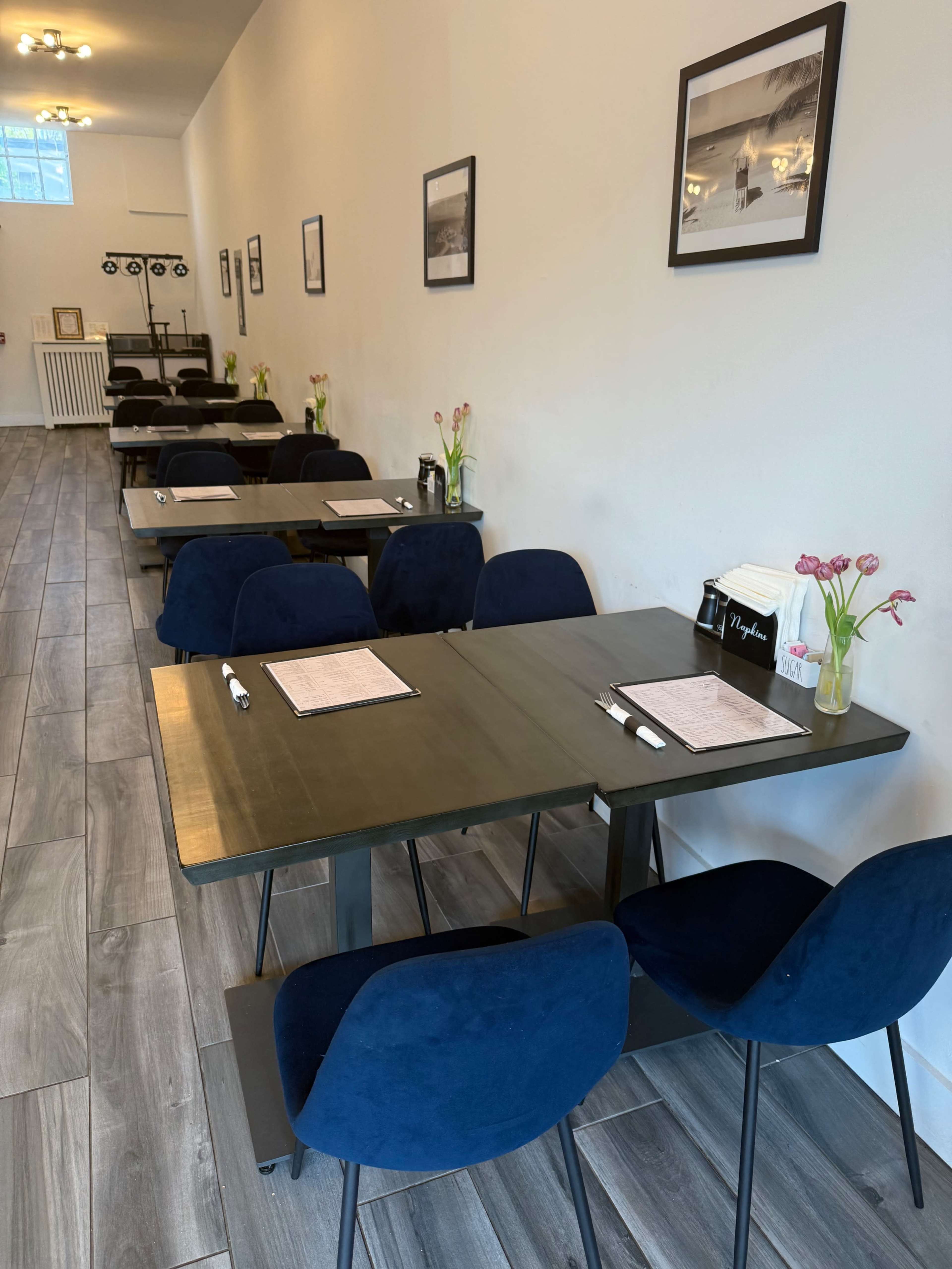 The image shows a row of tables with dark wood surfaces and navy blue chairs arranged neatly in a brightly lit interior space, featuring framed photographs on the walls.