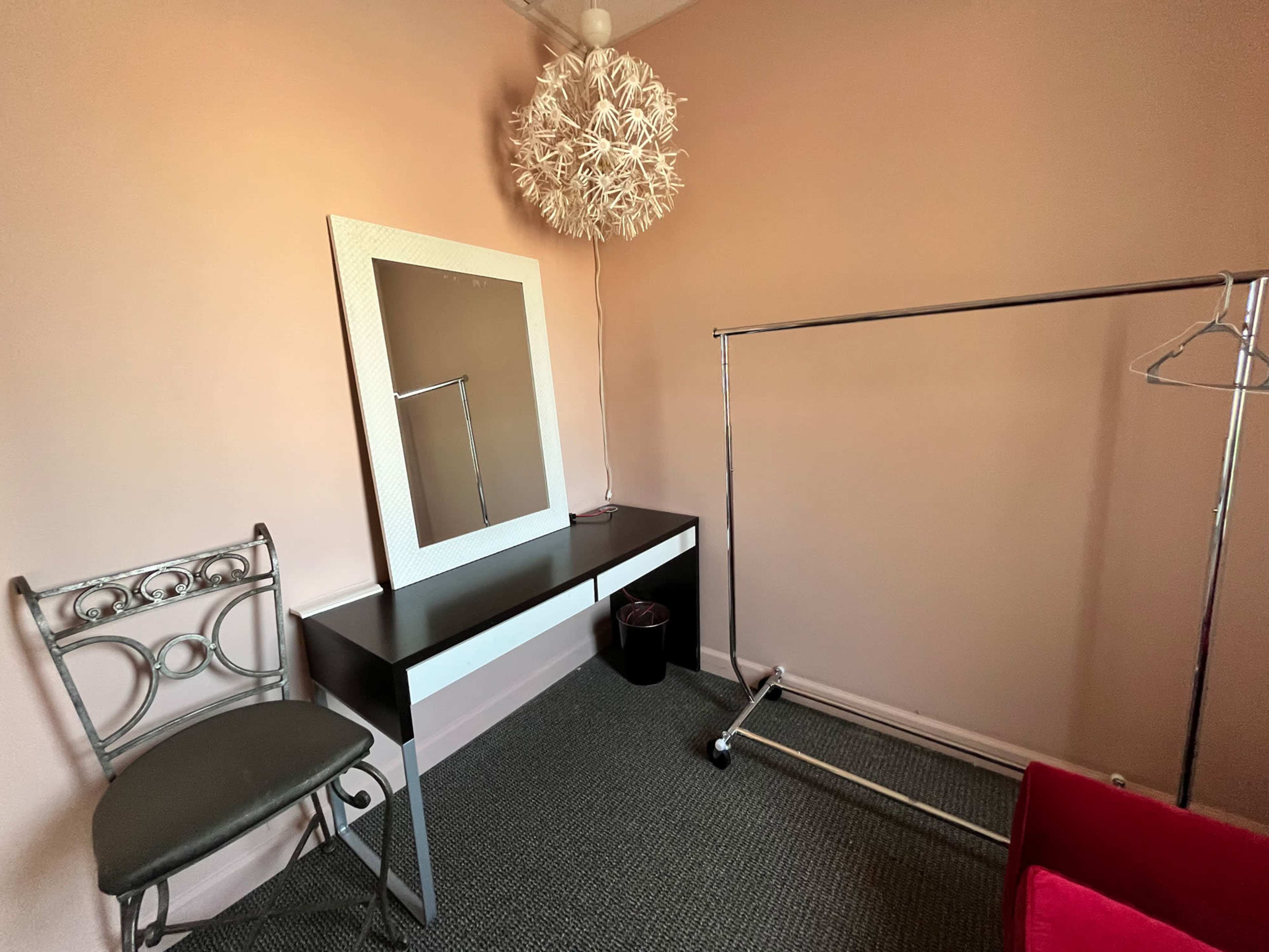 The interior of a small room features a black and white dresser with a mirror, a chair, a clothing rack, and a decorative light fixture.