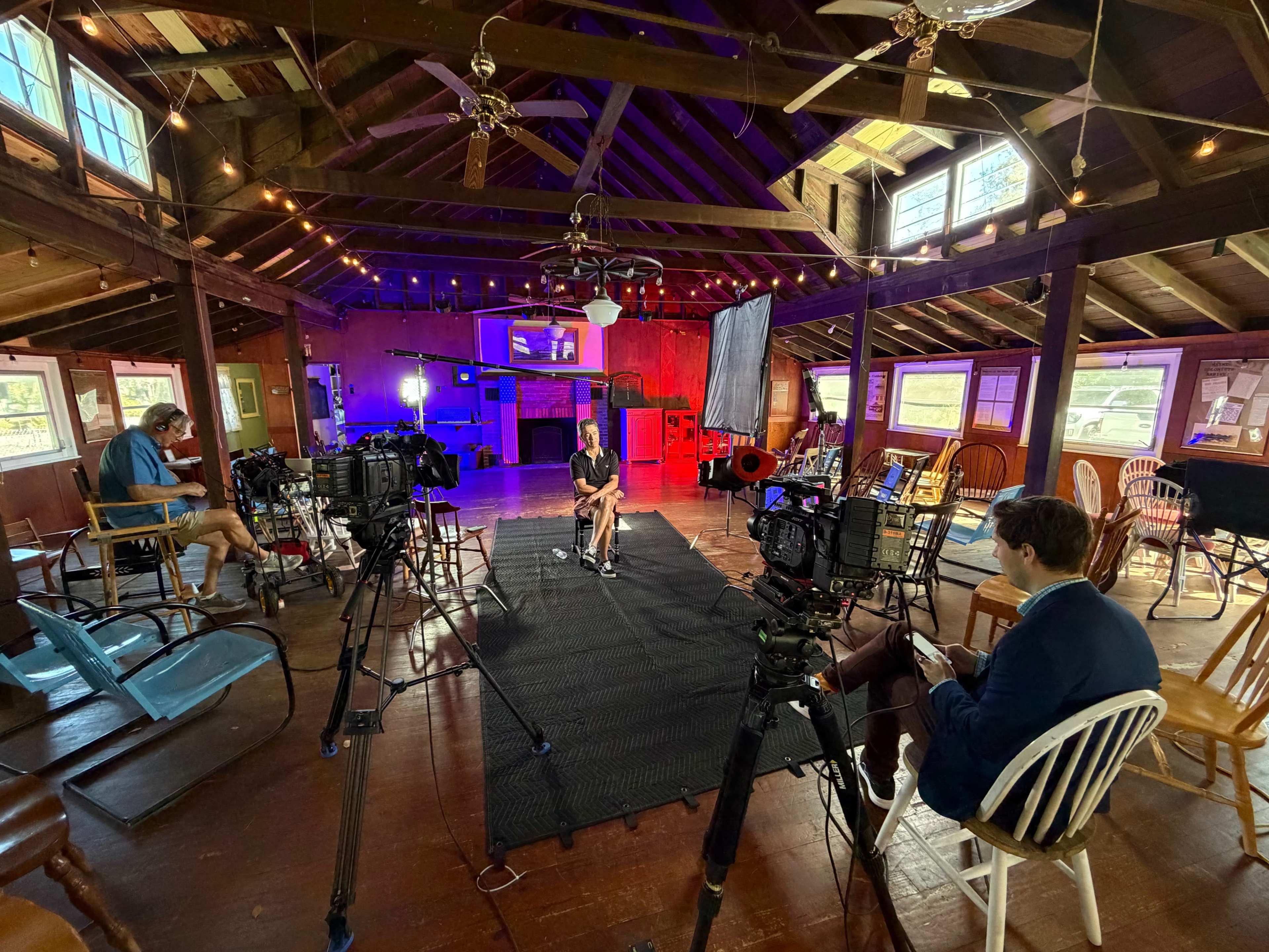 A film set is set up in a spacious, rustic room with wooden beams, featuring multiple cameras aimed at a person sitting on a mat in the center.