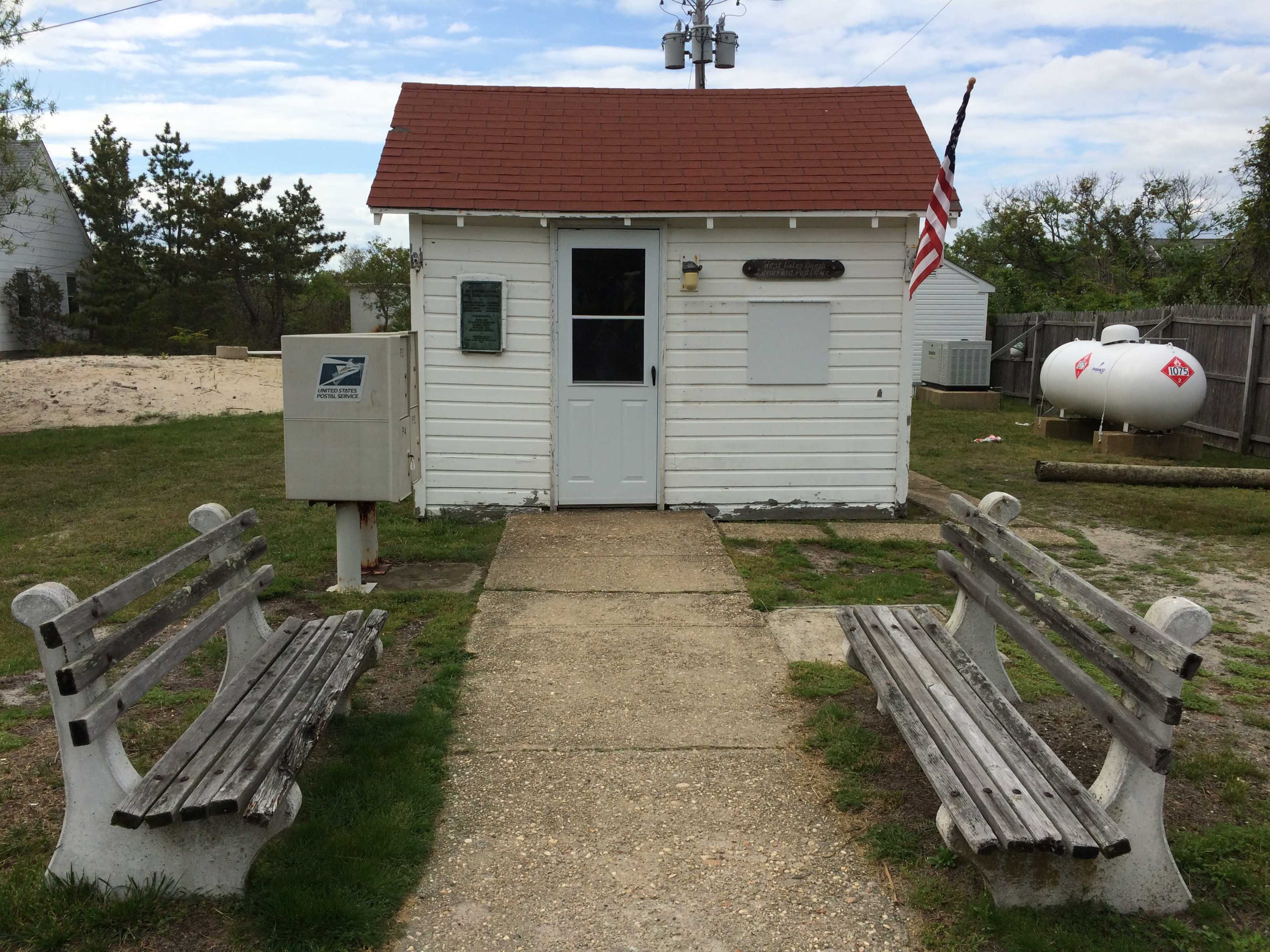 A small white building with a red roof and an American flag stands at the end of a concrete path, flanked by two wooden benches and a mailbox nearby.