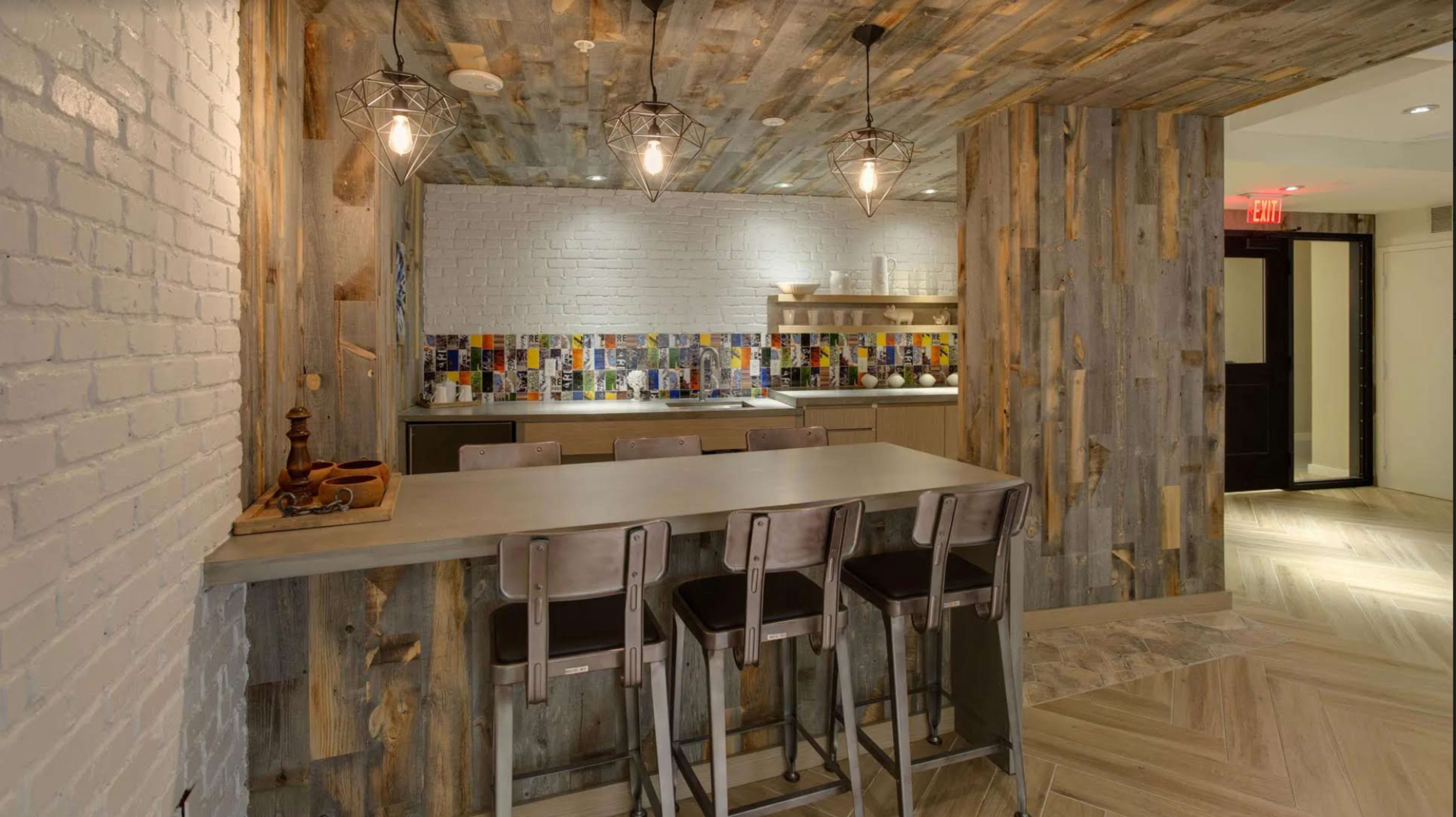 A modern kitchen features a wooden accent wall, a sleek bar with metal stools, and colorful tiled backsplash under warm pendant lighting.
