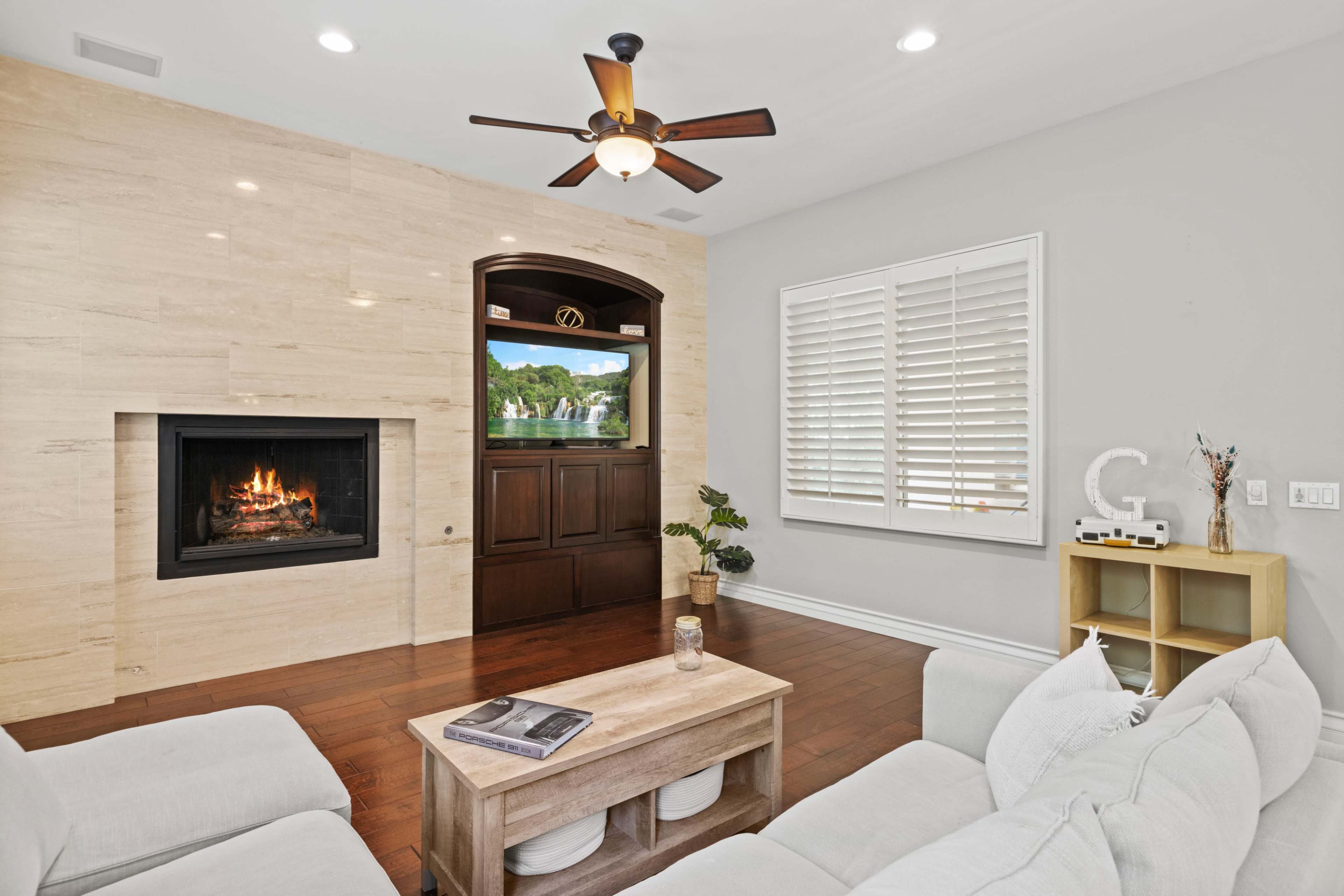 A cozy living room features a light-colored sectional sofa, a simple wooden coffee table, a fireplace, and a window with closed shutters.
