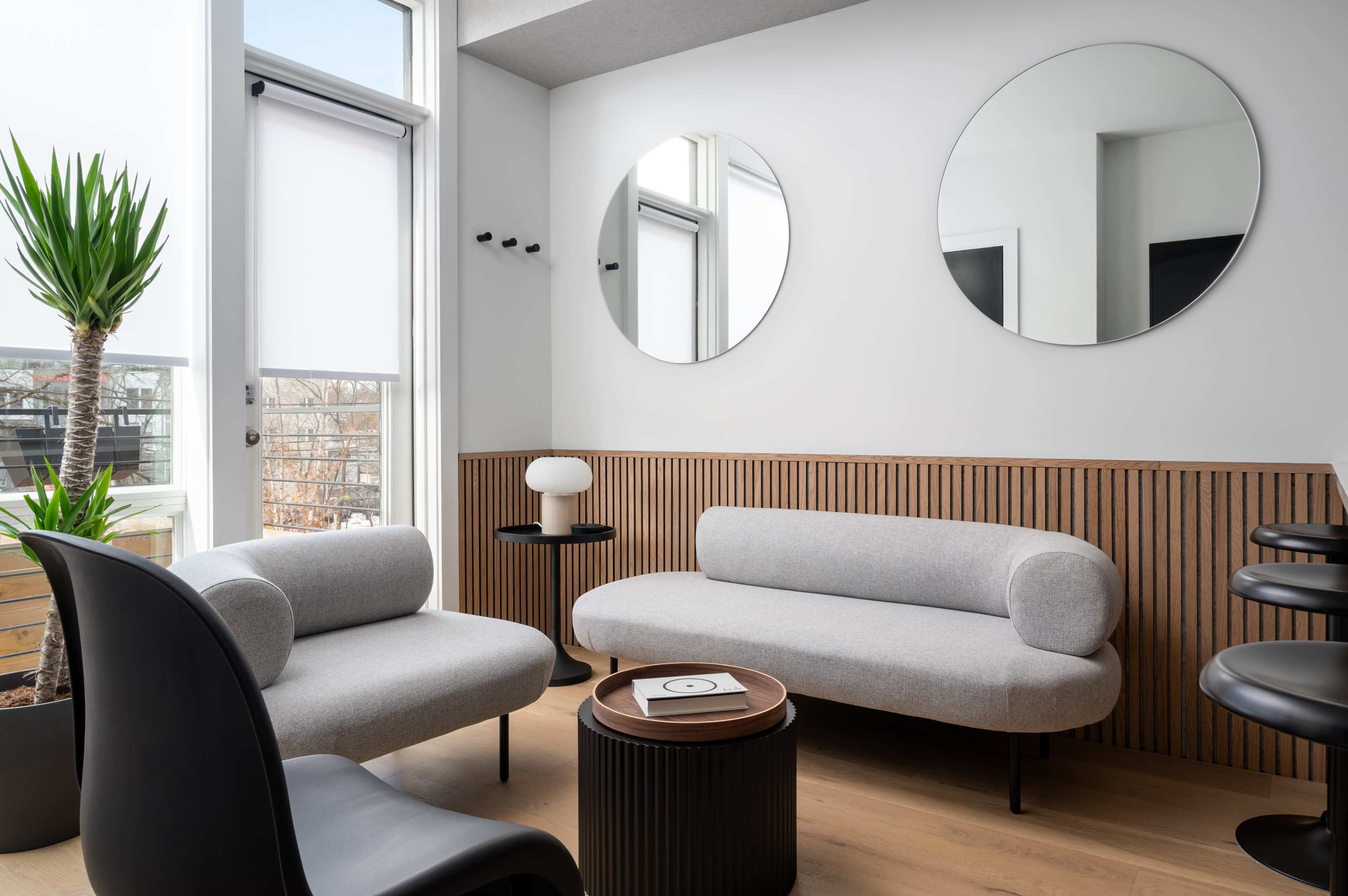 The image shows a modern lounge area with a light gray sofa, two circular mirrors on the wall, a black chair, and a round coffee table.