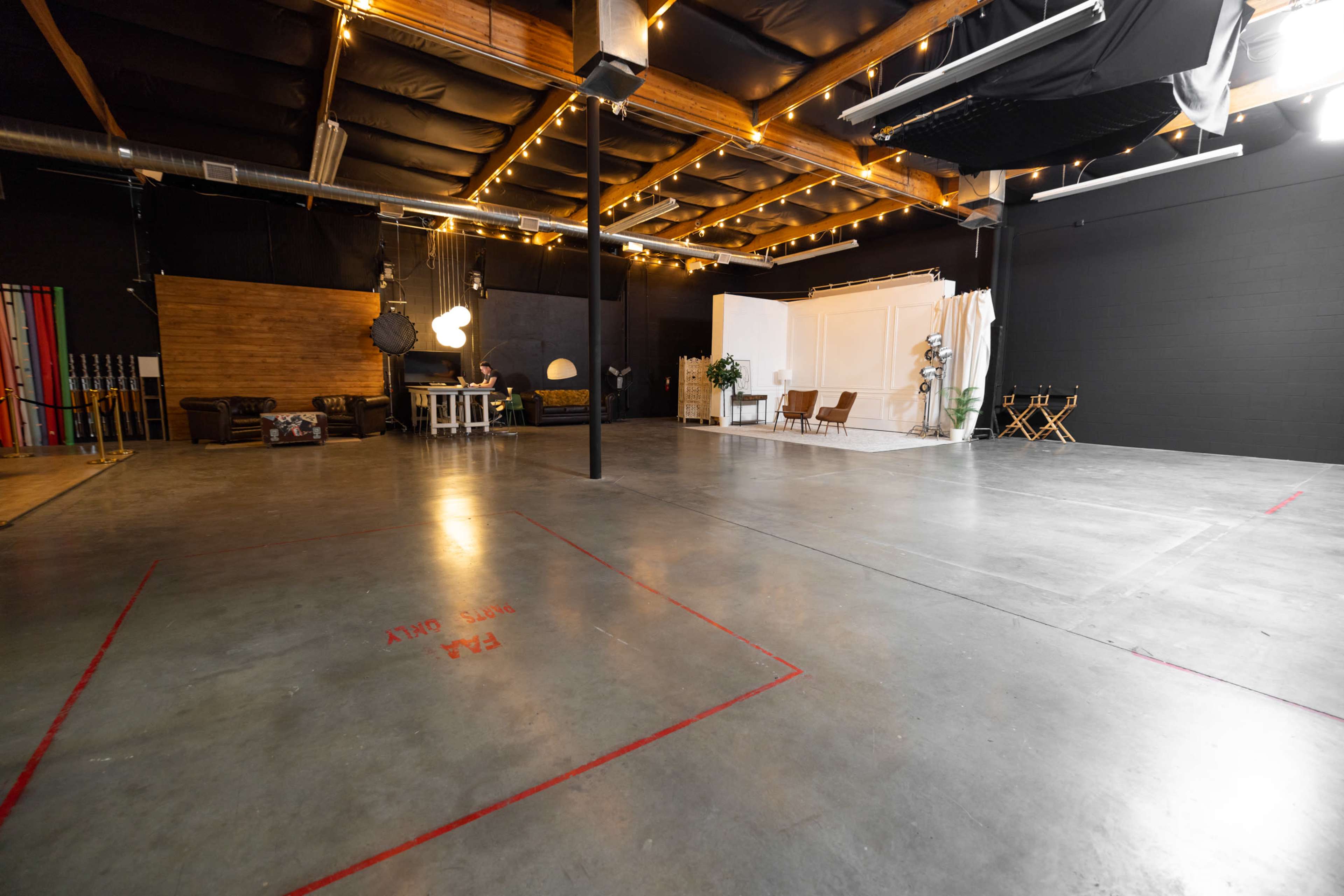 The image shows a large, empty studio space with concrete flooring, wooden beams, and various furniture set up on one side, including couches and a table.