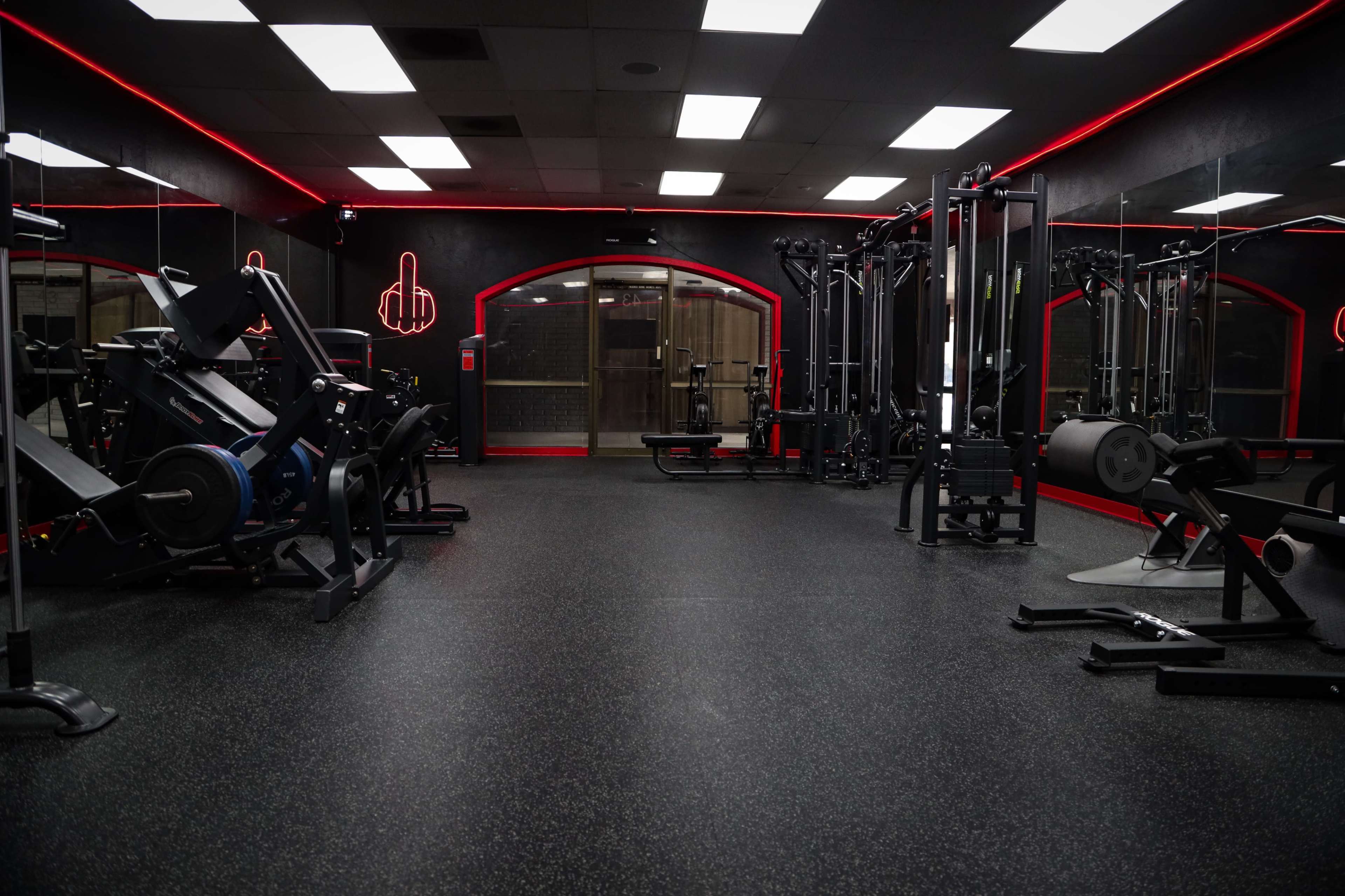 The image shows a dimly lit gym with various workout equipment arranged on a rubber floor and neon lighting along the walls.