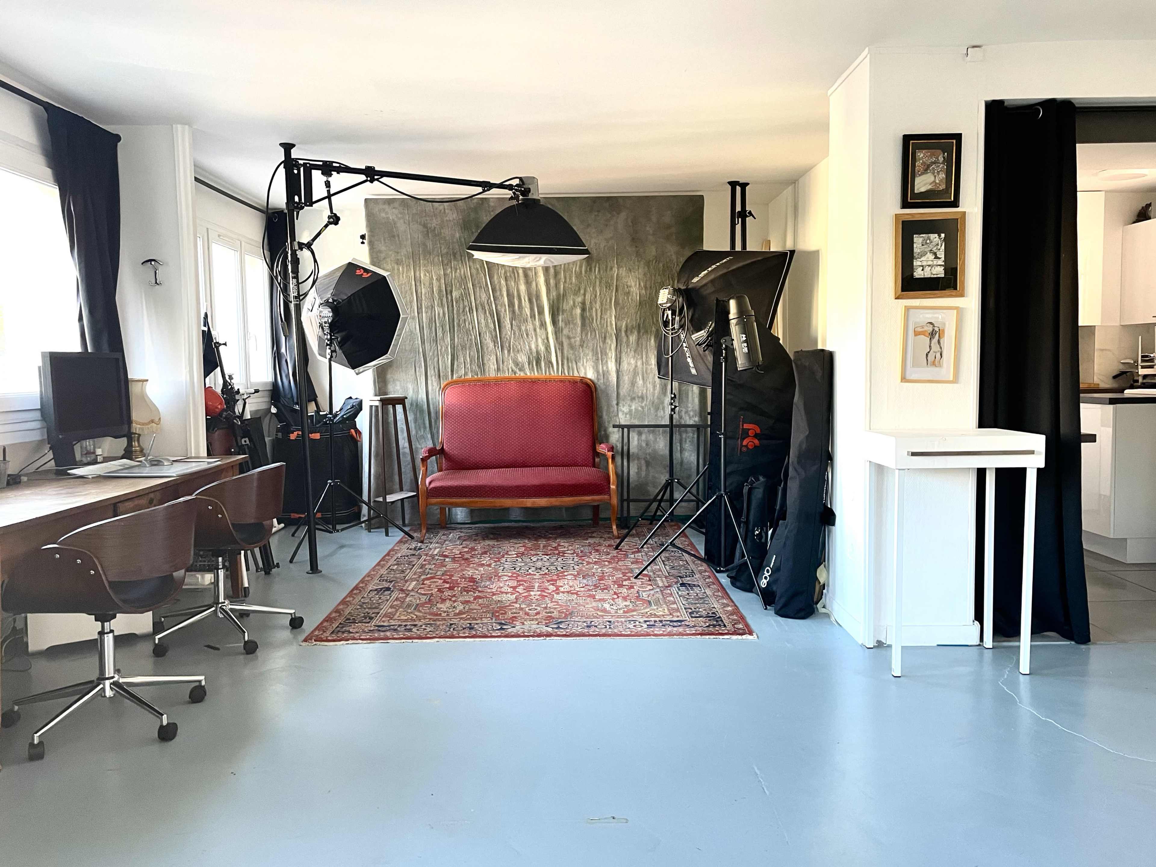 The image shows a photography studio with a red vintage sofa, lighting equipment, a desk with chairs, and a decorative rug on a concrete floor.
